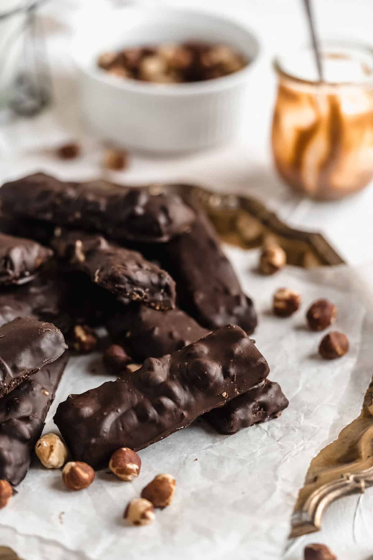 Vegan Hazelnut Snicker Bars are arranged on white parchment paper on a vintage silver tray. A jar of caramel sauce is in the background and additional hazelnuts are sprinkled around.