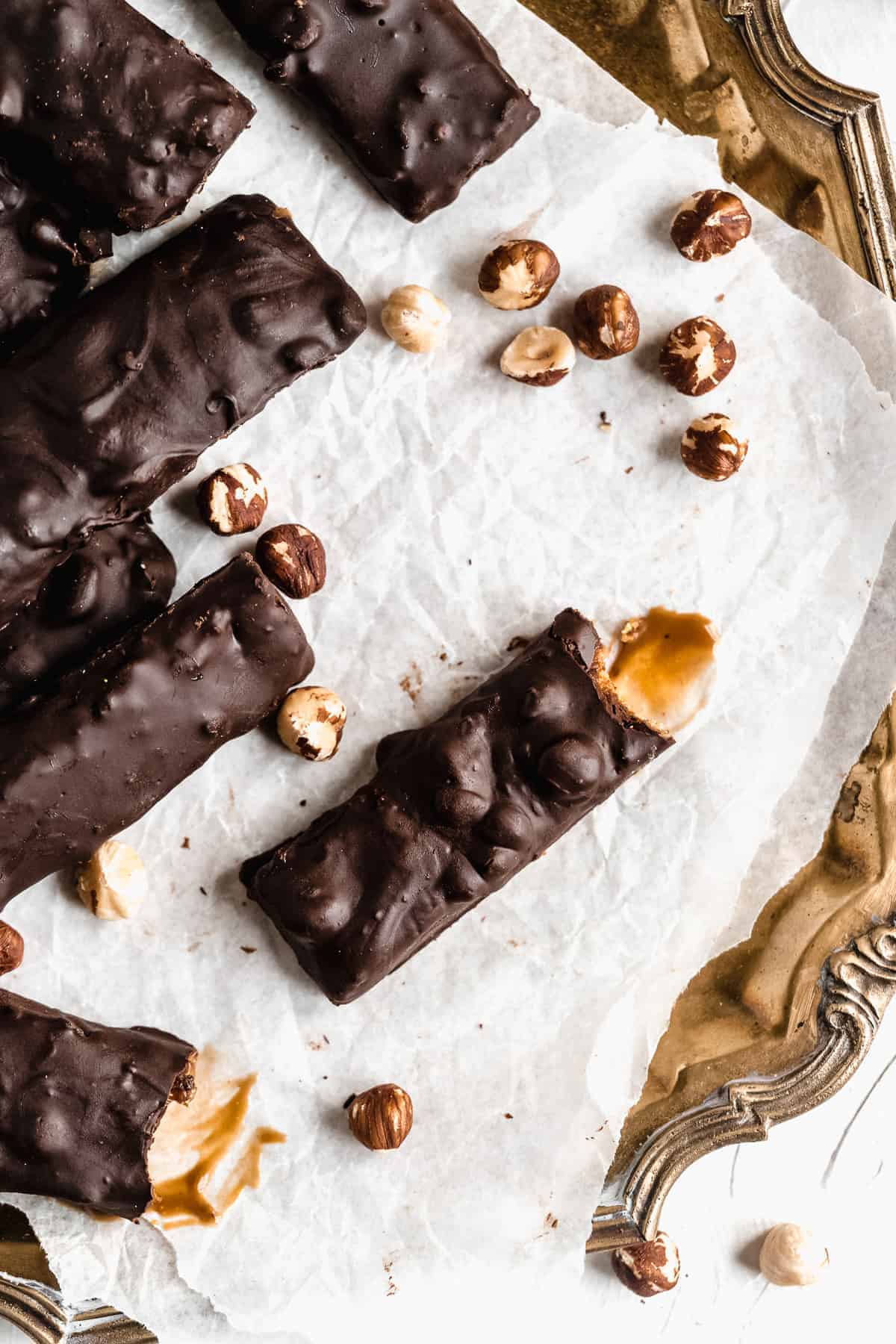 Overhead photo of Vegan Hazelnut Snicker Bars sitting on white parchment paper on a vintage silver tray.  Additional hazelnuts are sprinkled around.  