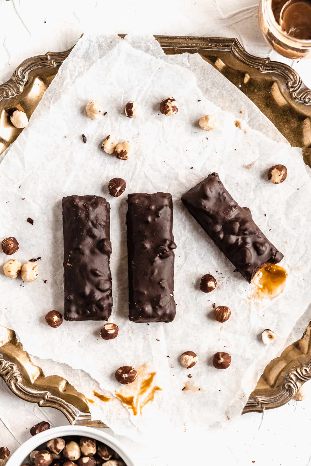 Overhead photo of three Vegan Hazelnut Snicker Bars sitting on white parchment paper on a vintage silver tray.  A bite is taken out of one bar to show the gooey inside.  Additional hazelnuts are sprinkled around.  