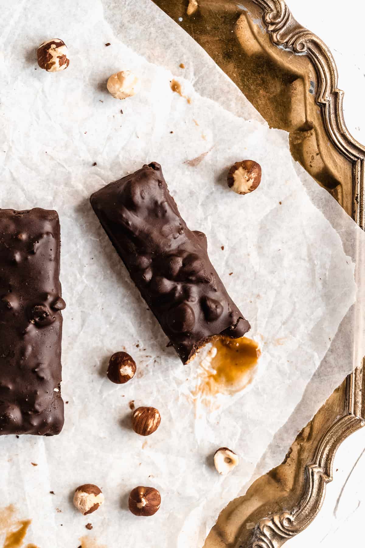 Overhead photo of two Vegan Hazelnut Snicker Bars sitting on parchment paper on a vintage silver tray.  A bite has been taken out of one bar.  Additional hazelnuts are sprinkled around.  