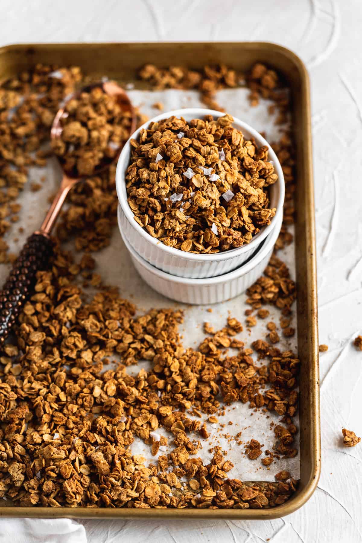 Overhead photo of a baking sheet with parchment paper and freshly baked Maple Sea Salt Nut-free Granola.  Two white ramekins are stacked and sitting on the baking sheet and a spoon has scooped up the granola and put it in the ramekins.  