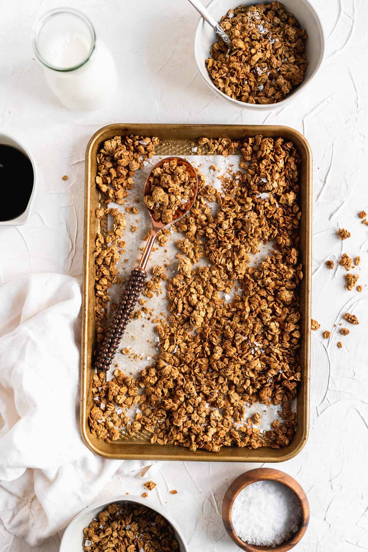 Overhead photo of freshly baked Maple Sea Salt Nut-free Granola on a baking sheet.  A white bowl is filled with granola.  A glass of milk and cup of coffee are placed nearby.  