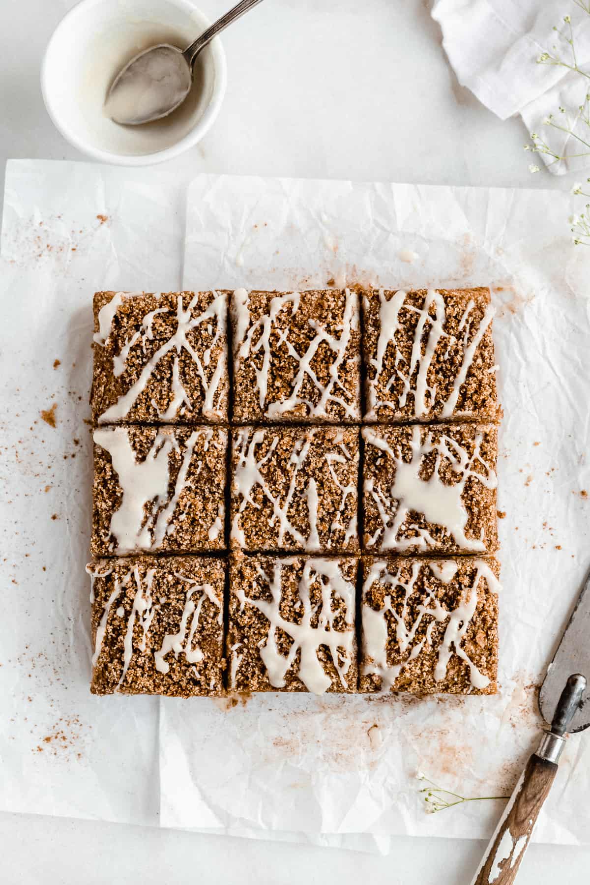 Overhead photo of freshly baked Paleo Almond Flour Coffee Cake cut into squares and sitting on parchment paper on a white marble slab. Cinnamon is sprinkled around the coffee cake.