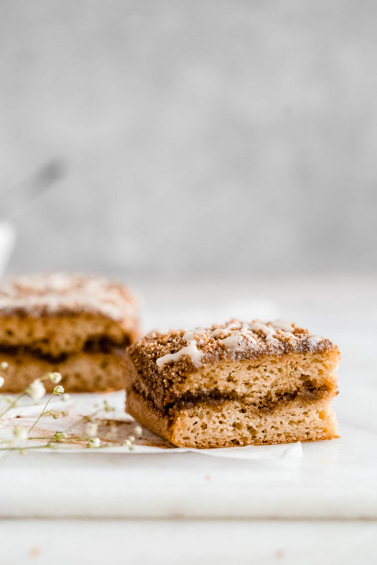 Side view closeup of a piece of Paleo Almond Flour Coffee Cake to reveal the texture of the cake and shows the cinnamon inside. The square piece of coffee cake is sitting on white parchment paper on a marble slab. Another piece has been placed in the background.