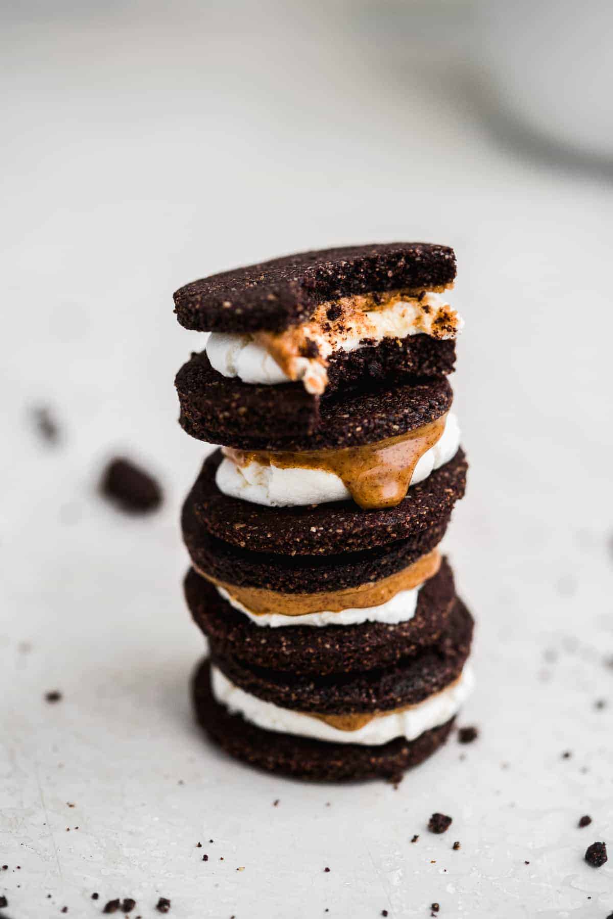 Close up side view photo of a stack of 4 Vegan Almond Butter Marshmallow Oreo Cookies.  The top cookie has a bite taken out of it.  Oreo cookie crumbs are sprinkled around.  