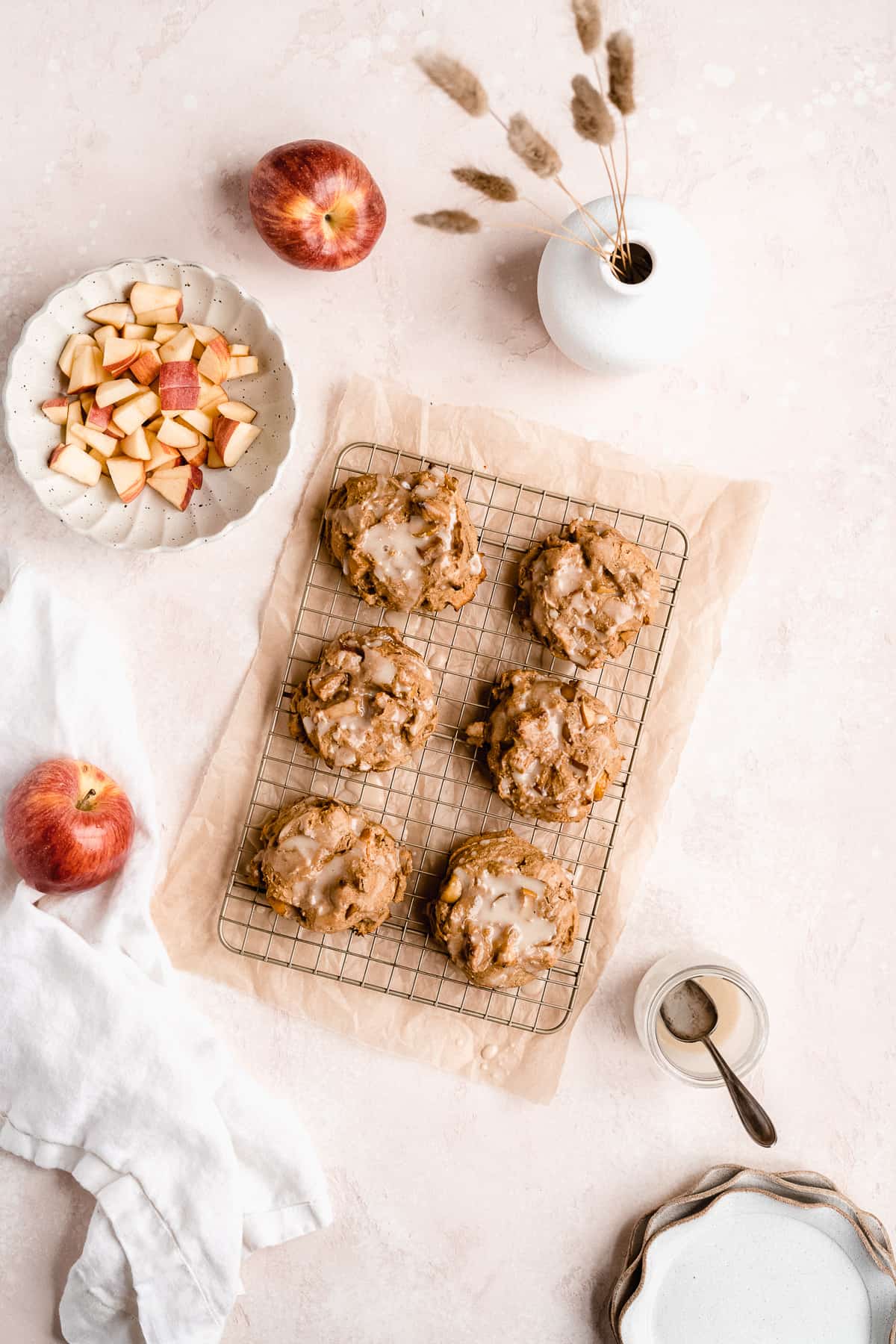 Overhead photo of freshly baked Vegan Baked Apple Fritters with Maple Coconut Butter Glaze sitting on a metal cooling rack on parchment paper.  Apple chunks in a white scalloped bowl and two whole apples are sitting nearby.  Three additional white scalloped plates are sitting nearby.  
