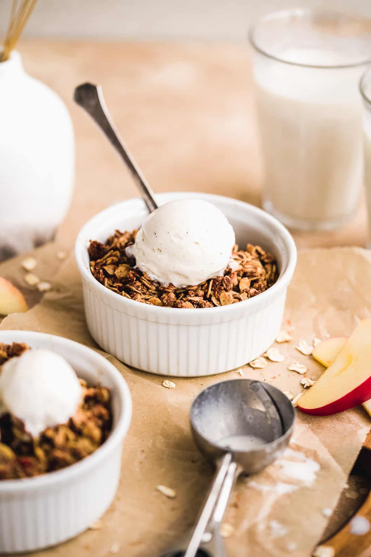 Side view photo of a white ramekin with  freshly baked apple crisp topped with vanilla ice cream.  An ice cream scoop, slices of apple and glass of milk are in the background.  