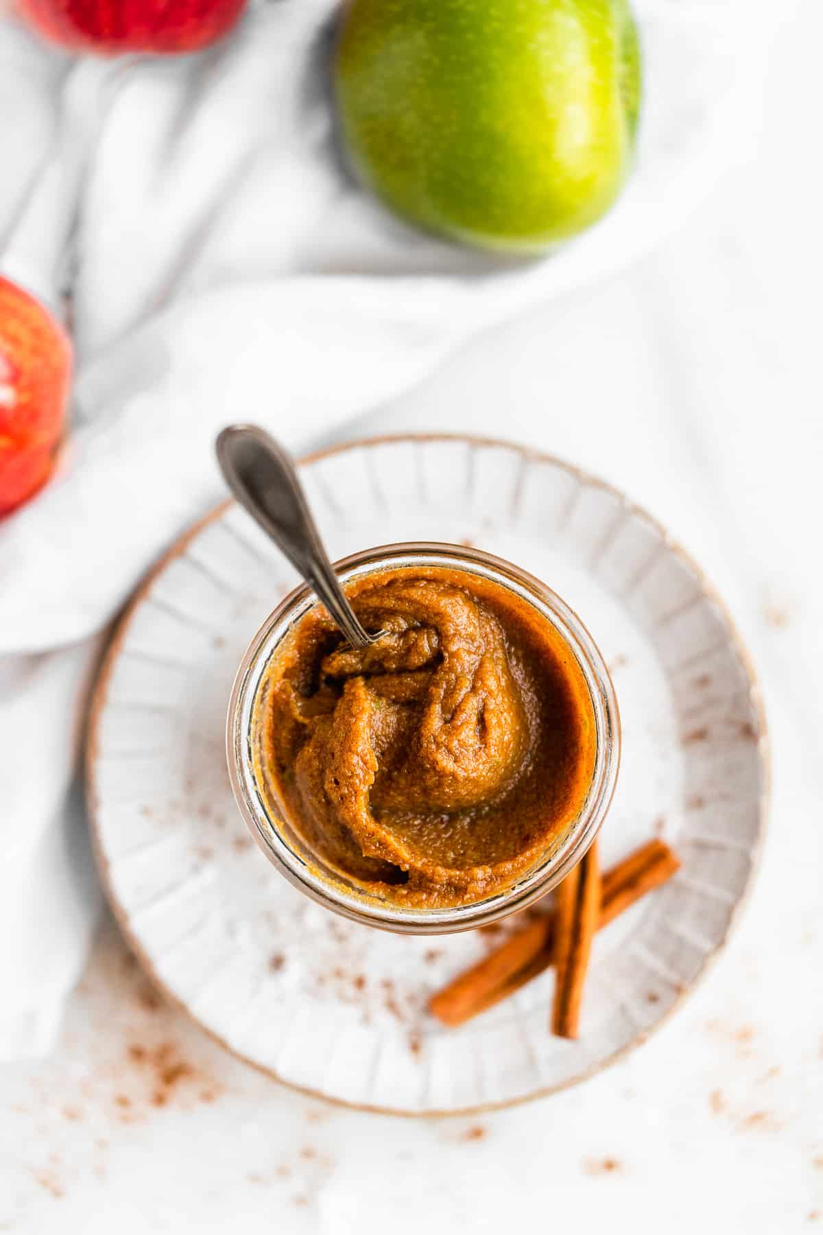 Overhead photo of a jar of Easy Salted Caramel Apple Butter sitting on a grey plate with a spoon standing up in the jar.  Two cinnamon sticks are sitting nearby and some green and red apples are in the background.  