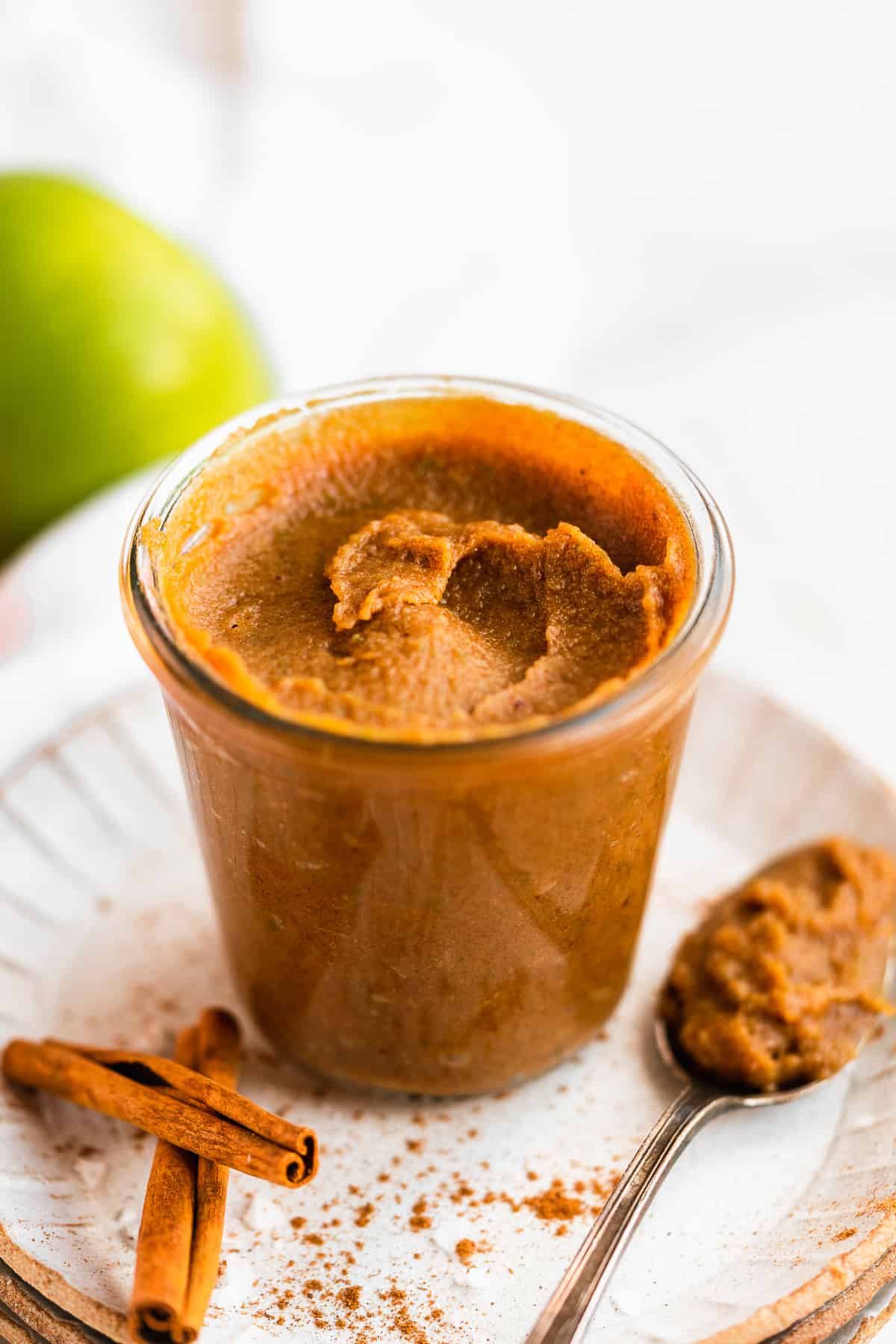 Clear jar sitting on a white plate and  filled to the top with Easy Salted Caramel Apple Butter.  A spoon with a scoop of Apple Butter is resting on the plate.  