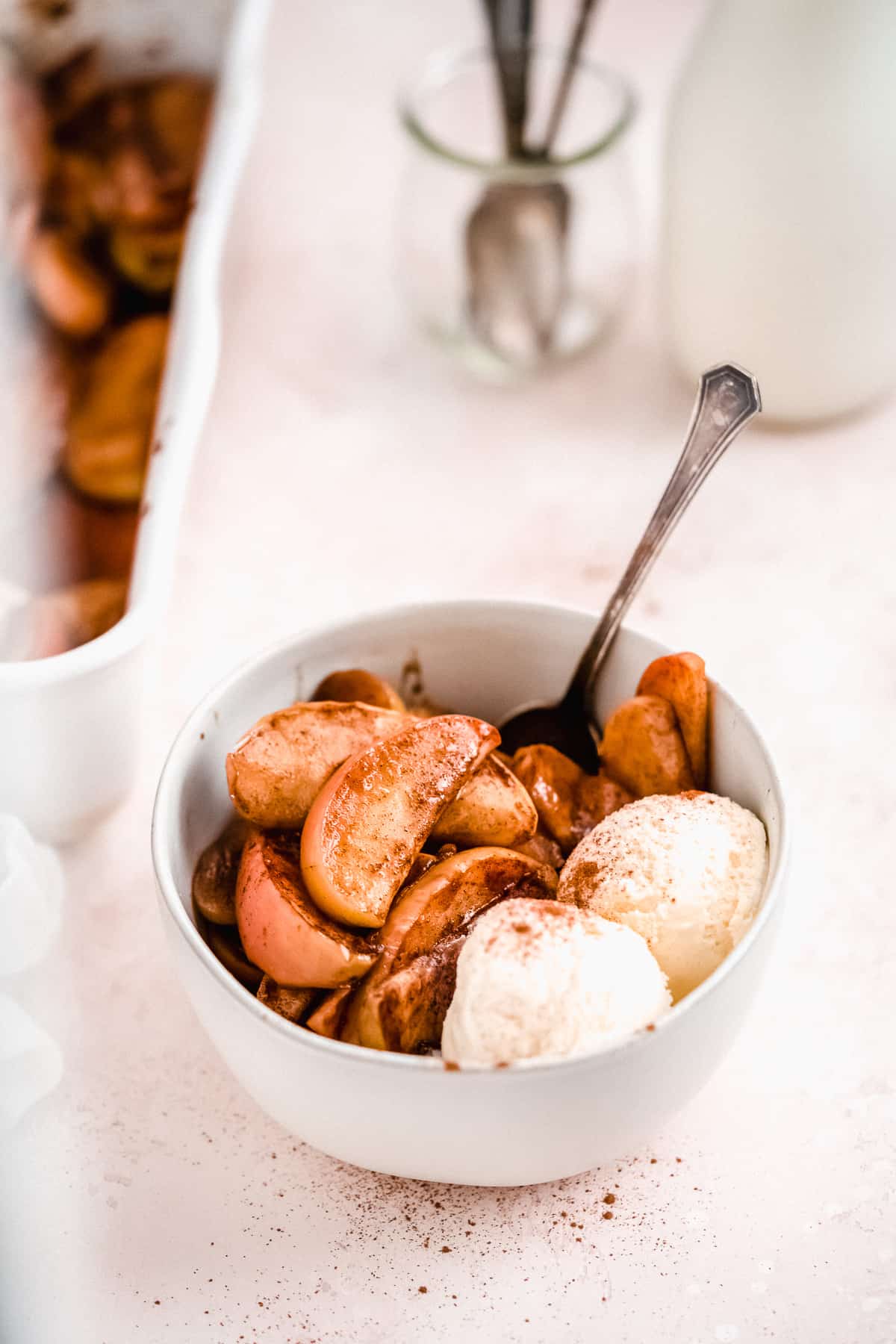 Side view photo of Easy Baked Cinnamon Apples in a white bowl with two scoops of vanilla ice cream and a spoon just waiting to be enjoyed.  