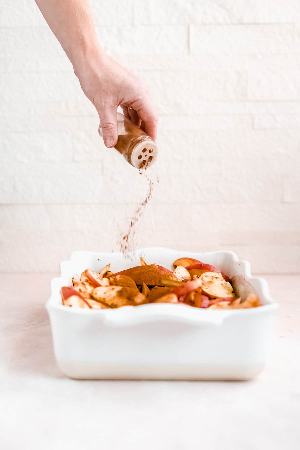 Picture of the recipe being made showing a white square scalloped baking dish with the sliced apples layered in the dish and cinnamon being sprinkled over them from above.  