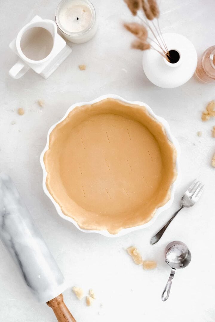 Overhead view of the completed pie crust ready to be baked. Fork pricks have been made in the bottom of the crust. A fork, measuring cup and rolling pin are placed nearby.