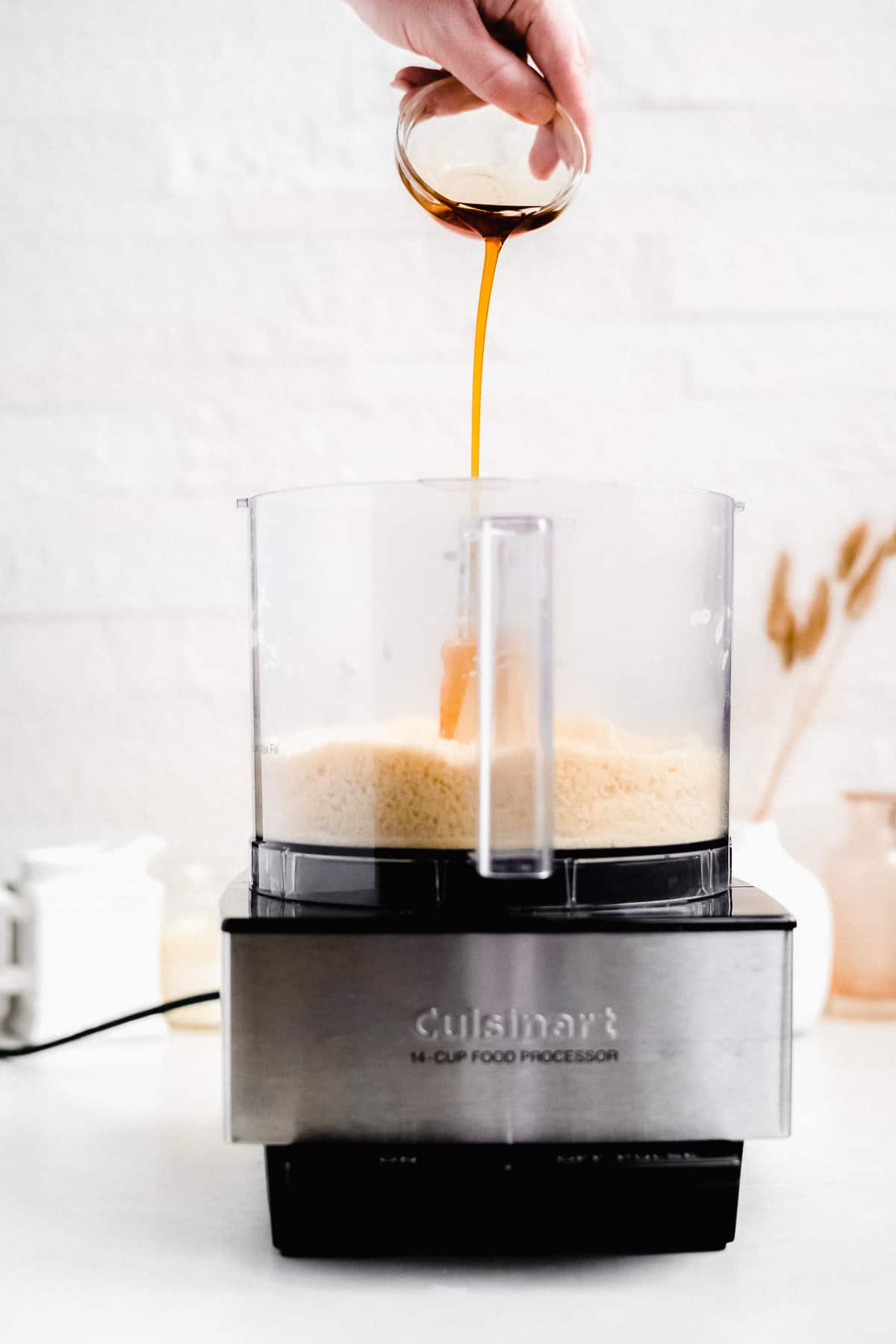 Side view of the food processor during the process of making the crust. Dry ingredients are in the bottom of the food processor and the coconut oil is being poured in from above.