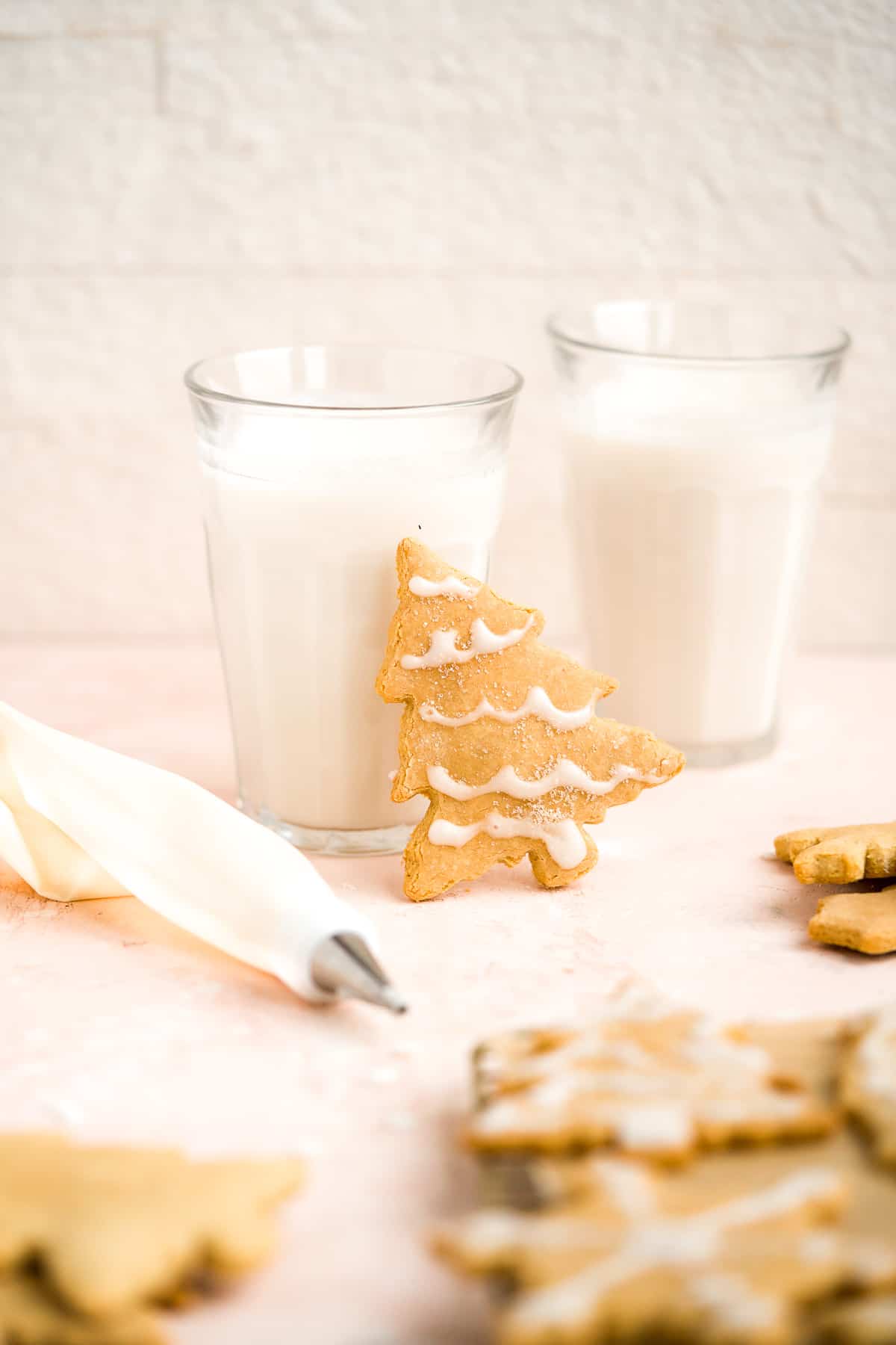 A single Soft and Crispy Gluten-free Sugar Cookie leaning against two clear glasses of milk. Additional cookies are arranged around the glasses.