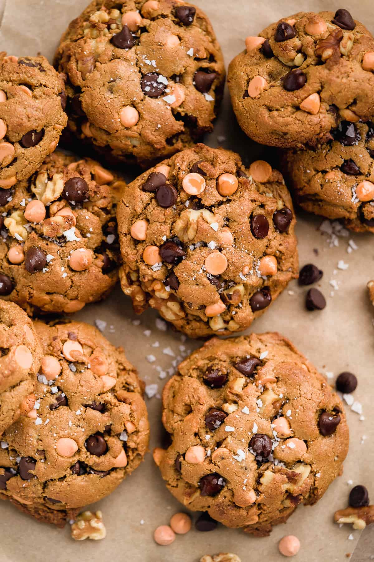 Closeup photo of 8-9 butterscotch chocolate chip cookies scattered on parchment paper. Chocolate chips and sea salt are sprinkled nearby.