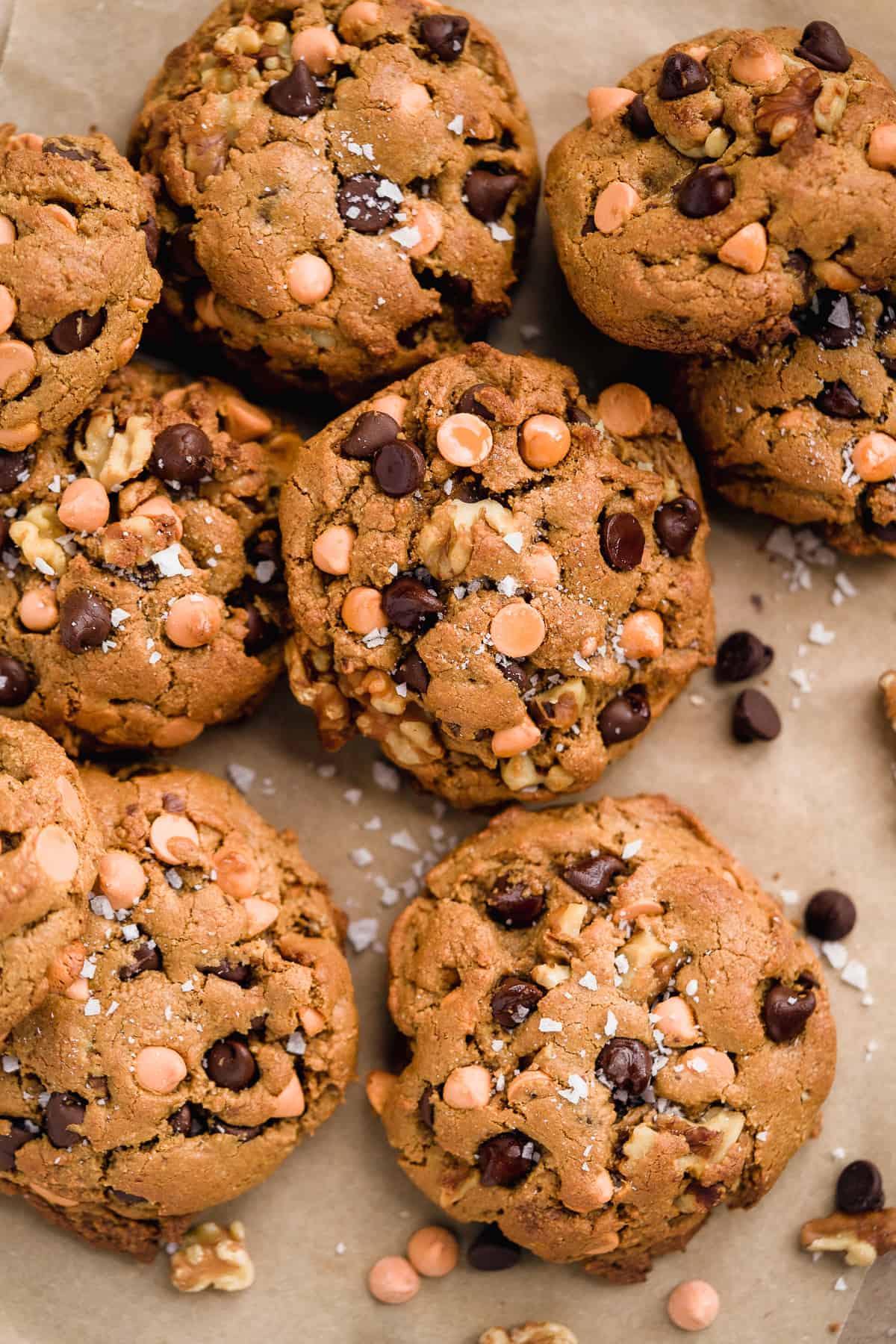 Closeup photo of 8-9 butterscotch chocolate chip cookies scattered on parchment paper.  Chocolate chips and sea salt are sprinkled nearby.  