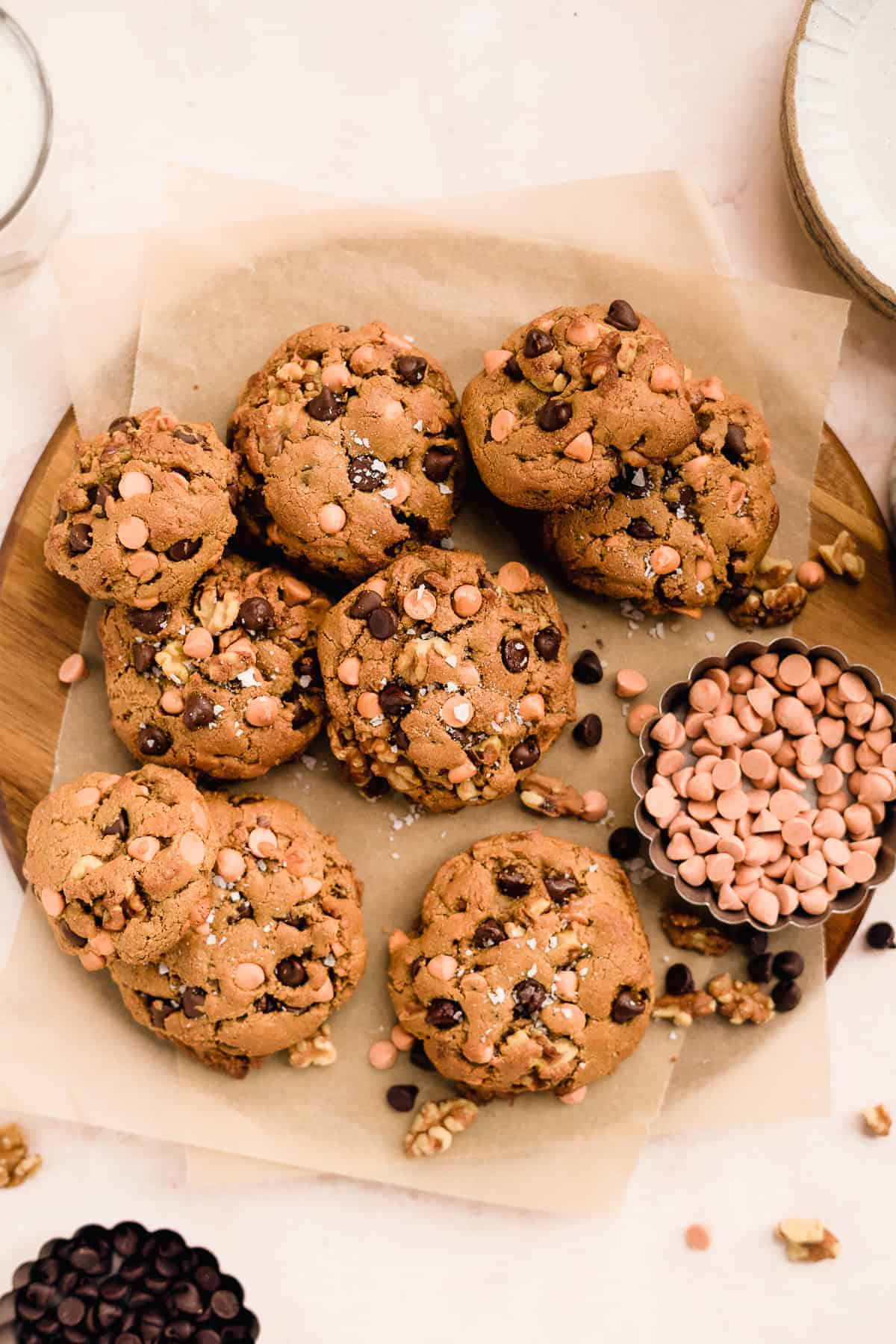 Nine yummy butterscotch chocolate chip cookies spread out on parchment paper on top of a round wood platter. Small scalloped bowl with butterscotch chips are nearby. Chocolate chips and walnuts are sprinkled around.