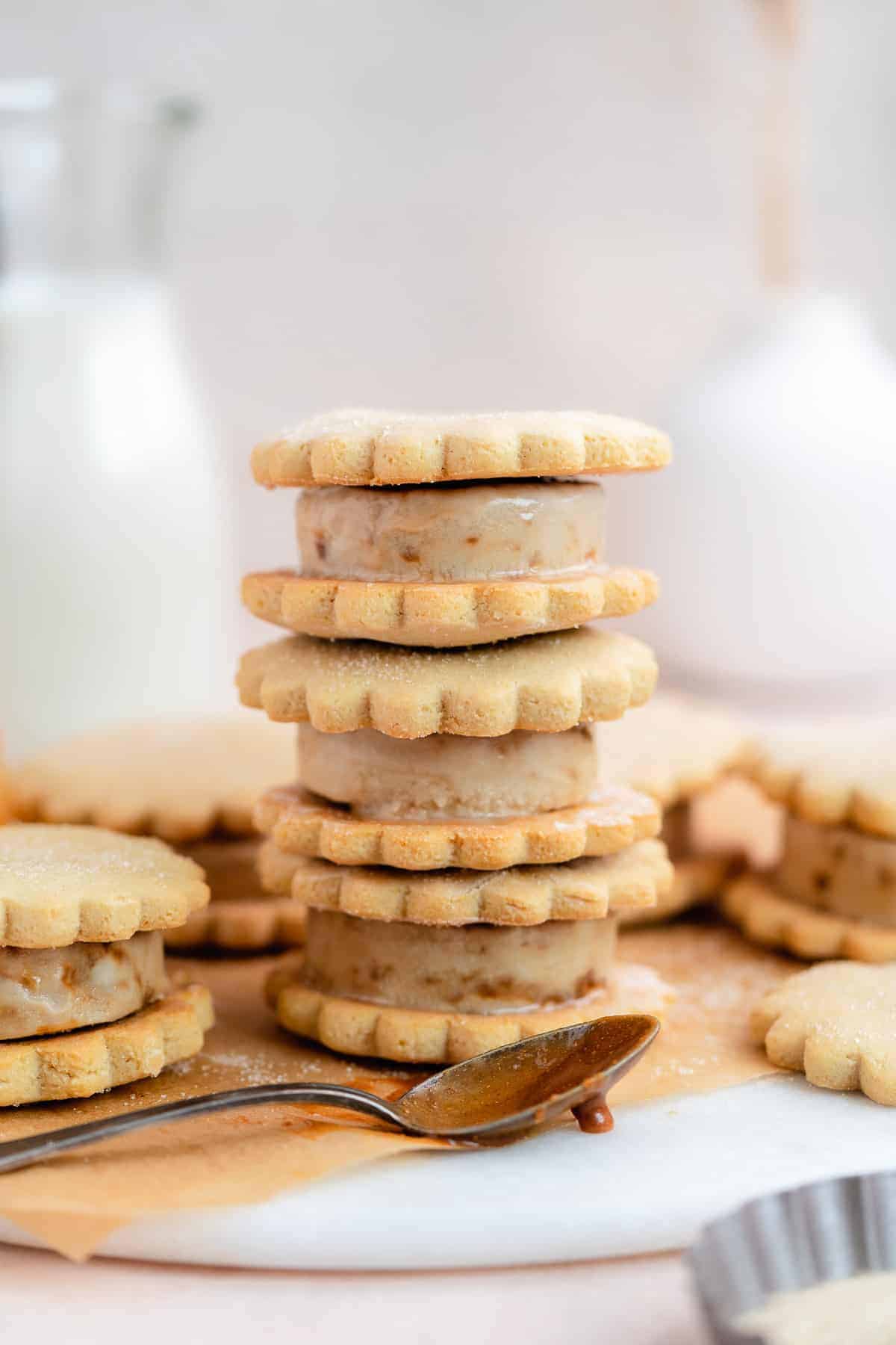 Side view of stack of 3 caramel sugar cookie ice cream sandwiches on parchment paper and marble slab.  One ice cream sandwich and silver spoon dipped in caramel sauce are resting nearby.  