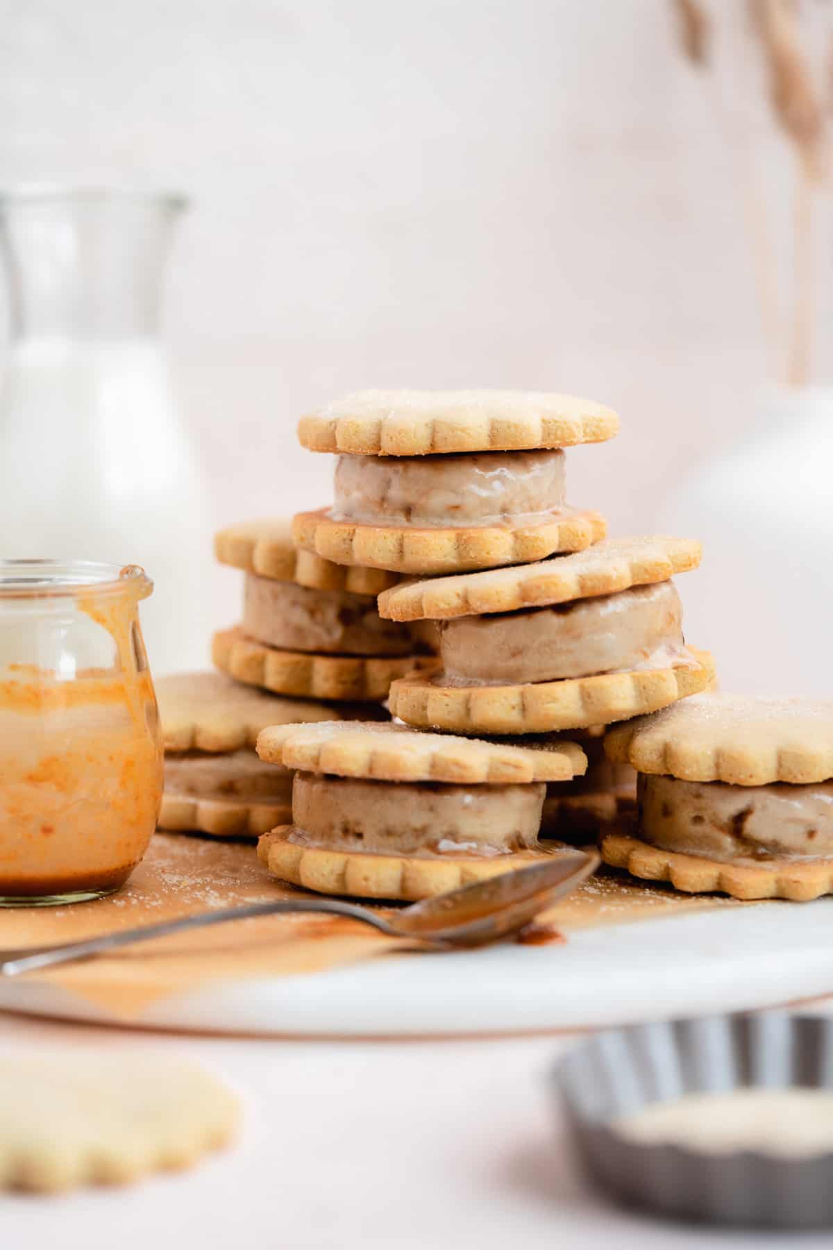 Side view photo of 7 ice cream sandwiches stacked in a pyramid sit on top of parchment paper on a marble slab.   Small clear jar with caramel sauce and silver spoon dipped in caramel sauce are placed nearby.  