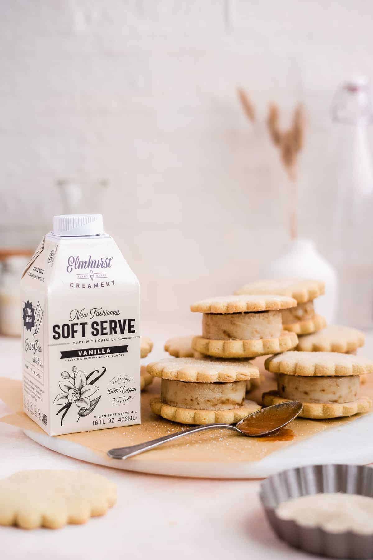 Photo shot from a the side showing multiple stacks of caramel sugar cookie ice cream sandwiches sitting on parchment paper on top of a marble slab.  A carton of Elmhurst's new dairy free ice cream mix and a silver spoon dipped in caramel sauce are placed nearby.  