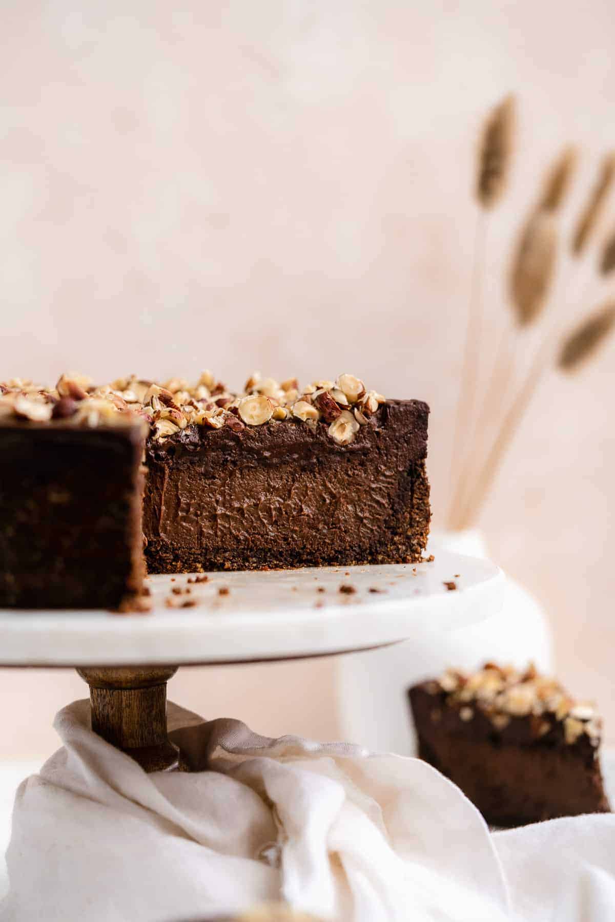 Side view close up of Chocolate Toasted Hazelnut Cheesecake sitting on top of a  white marble cake plate.  A section of the cheesecake is missing to reveal the delicious inside of this decadent dessert.  