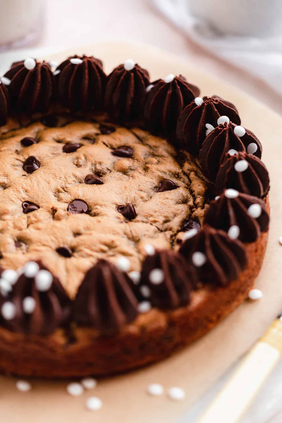 Close up photo of chocolate chip cookie cake on parchment paper.  Chocolate frosting piped around the edges of the cake.  