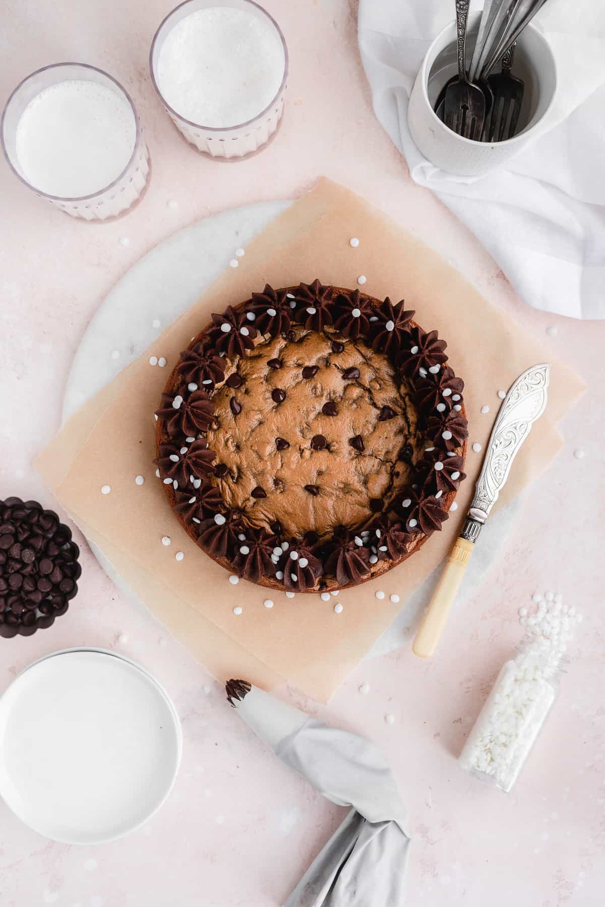Overhead view of chocolate chip cookie cake sitting on parchment paper and a marble slab.  Decorative knife is resting beside the cake.   Two glasses of milk, cup with multiple spoons, small dessert plates and bowl of chocolate chips are nearby.  