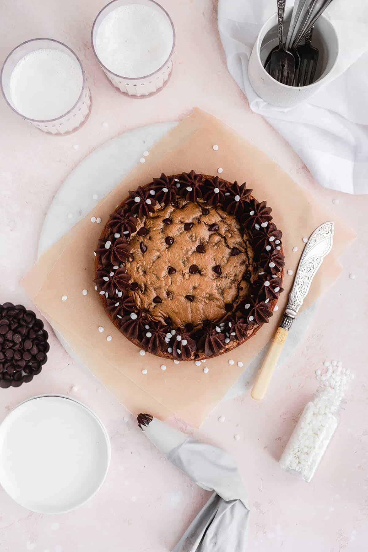 Overhead view of chocolate chip cookie cake sitting on parchment paper and a marble slab. Decorative knife is resting beside the cake. Two glasses of milk, cup with multiple spoons, small dessert plates and bowl of chocolate chips are nearby.