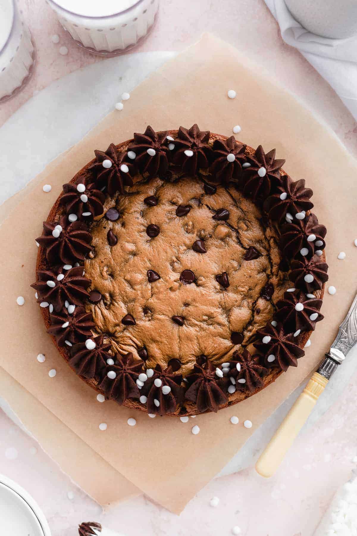 Overhead photo of round chocolate chip cookie cake with chocolate frosting piped around the edges. Silver knife with pearl handle is resting nearby. Cake is sitting on parchment paper on a white marble slab.