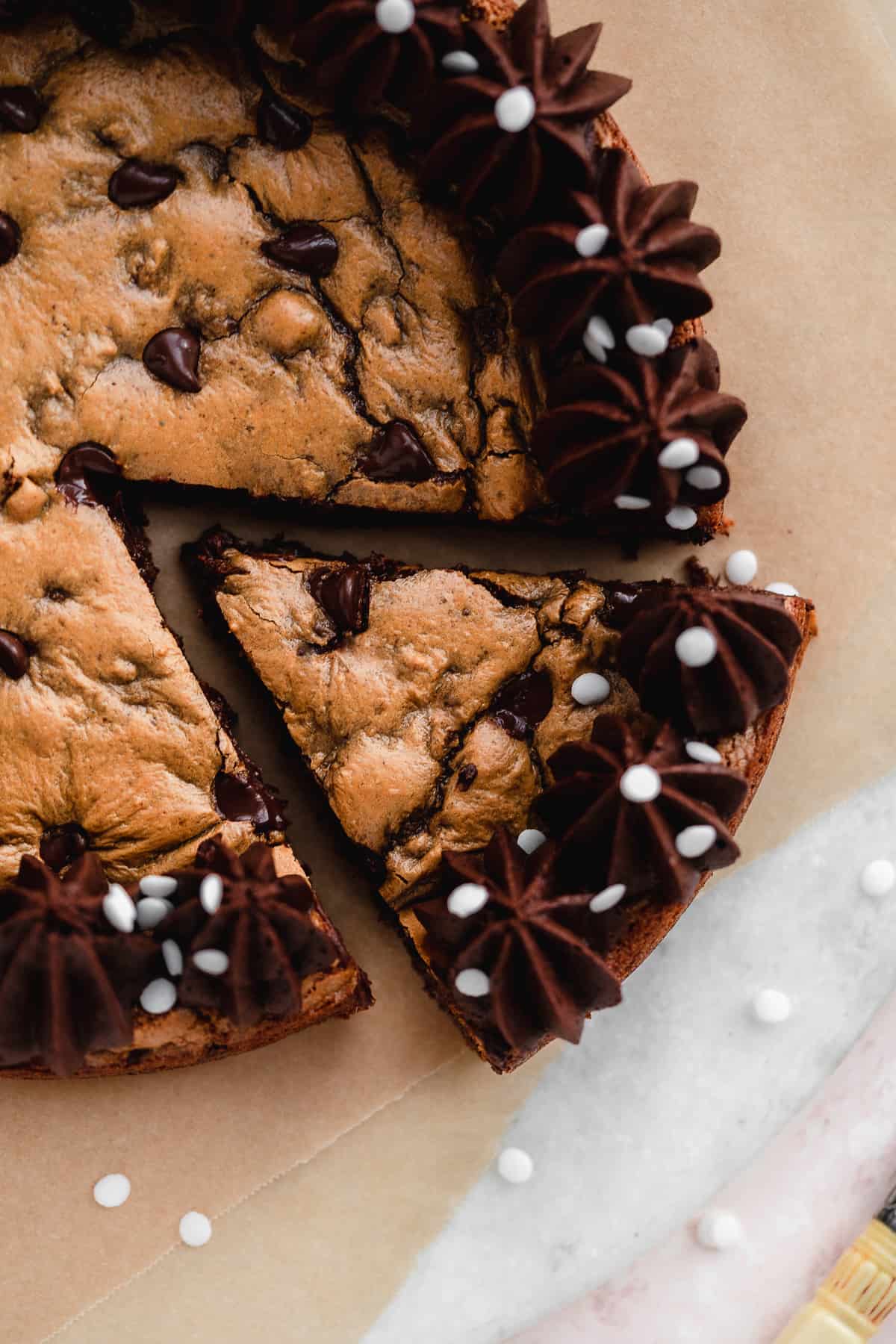Close up photo of chocolate chip cookie cake on brown parchment paper.  A triangular slice of cookie cake has been cut and placed nearby.  