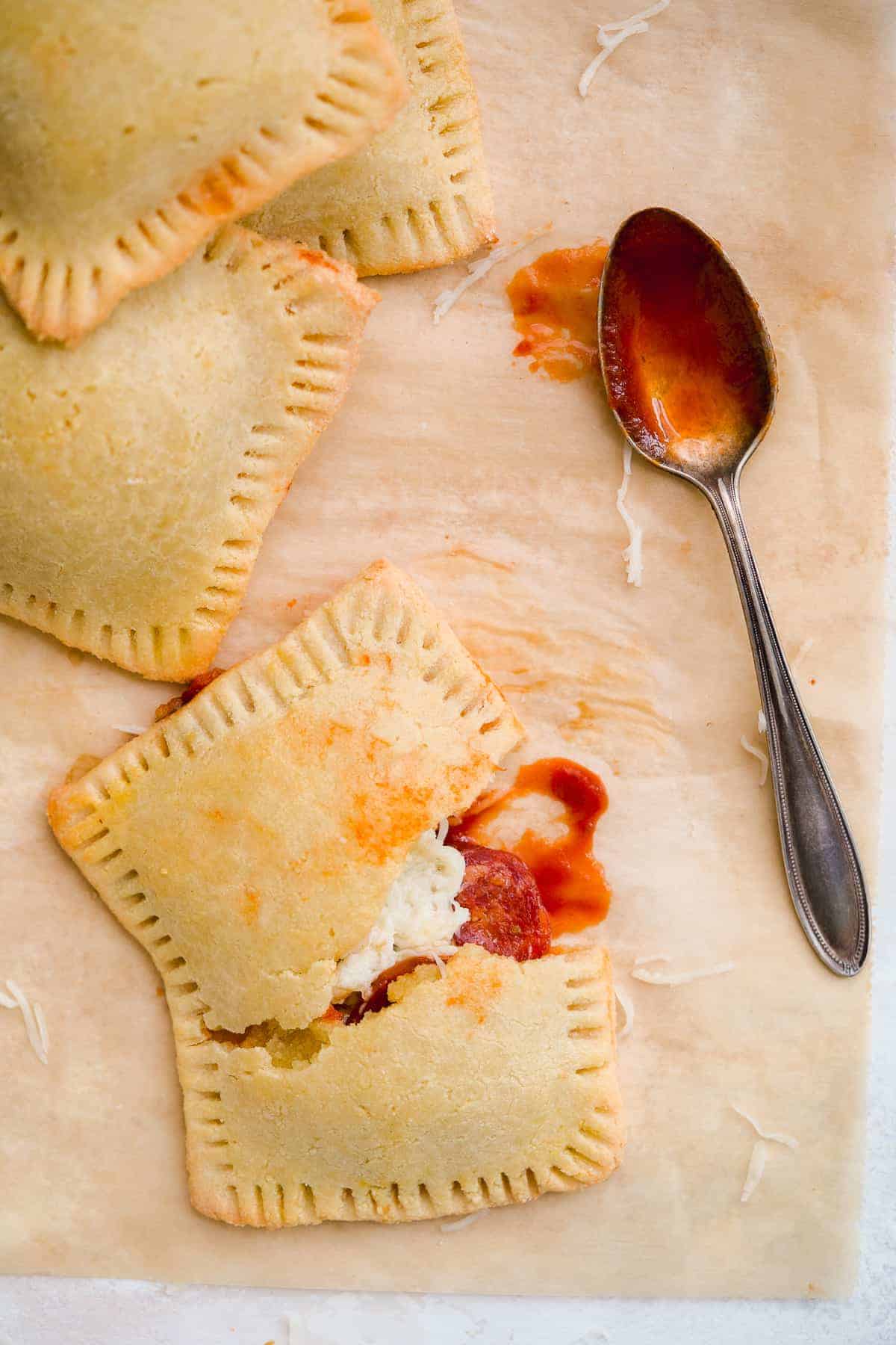 Parchment paper with several pizza hot pockets stacked.  One hot pocket is broken in half to reveal yummy filling inside.  Silver spoon dipped in marinara is resting nearby.  Shredded parmesan cheese is sprinkled around.  