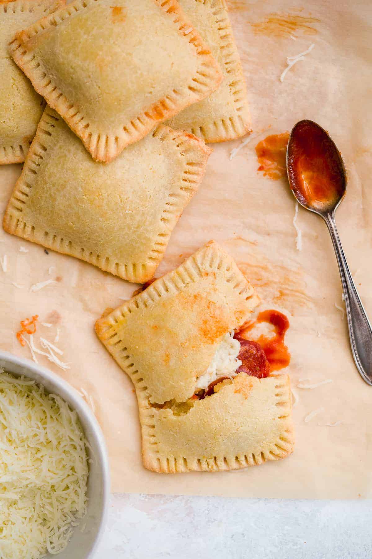 Four yummy pizza hot pockets scattered on brown parchment paper with a bowl of shredded parmesan cheese. One hot pocket is cut in half to reveal delicious insides. Silver spoon with marinara sauce in resting nearby.