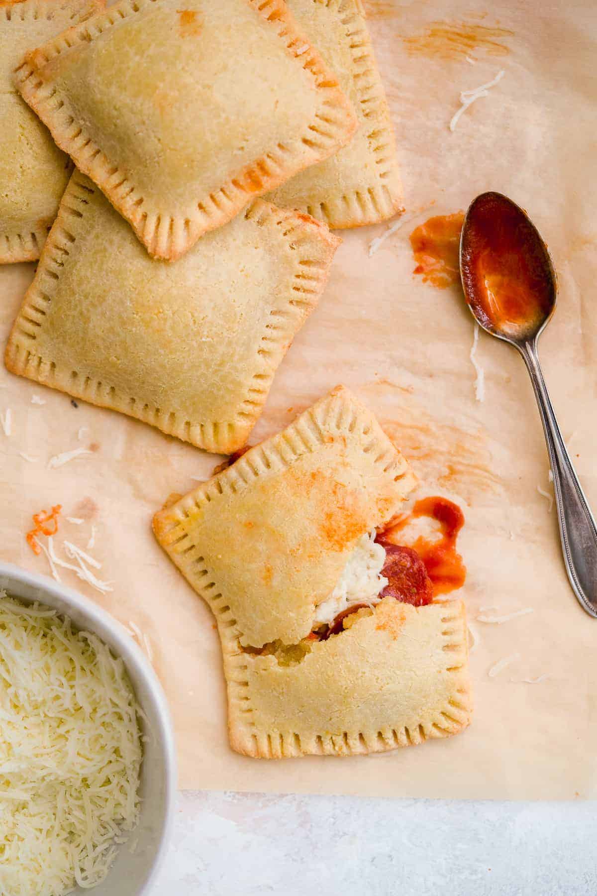 Four yummy pizza hot pockets scattered on brown parchment paper with a bowl of shredded parmesan cheese.  One hot pocket is cut in half to reveal delicious insides.  Silver spoon with marinara sauce in resting nearby.  