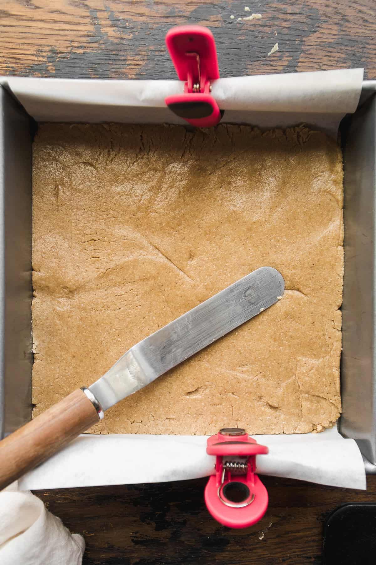 A baking pan lined with parchment paper holds an even layer of light brown vegan 7 layer bars dough. A metal spatula with a wooden handle rests on the dough, and two red clips secure the paper to the pan&rsquo;s edges.