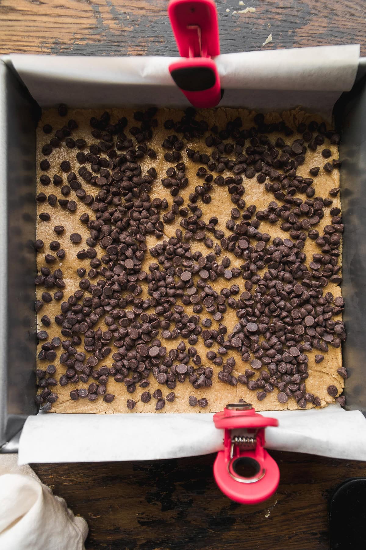 A square baking pan lined with parchment paper holds unbaked vegan 7 layer bars dough, topped with scattered chocolate chips and secured at two sides with red clips, on a wooden surface.