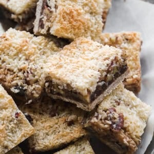 A close-up of a platter filled with vegan 7 layer bars. The bars feature a golden, toasted coconut topping, a middle layer of chocolate and nuts, and a cookie-like crust on the bottom.