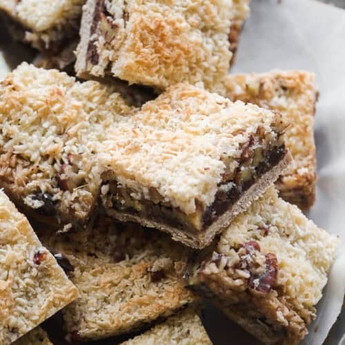 A close-up of a platter filled with vegan 7 layer bars. The bars feature a golden, toasted coconut topping, a middle layer of chocolate and nuts, and a cookie-like crust on the bottom.