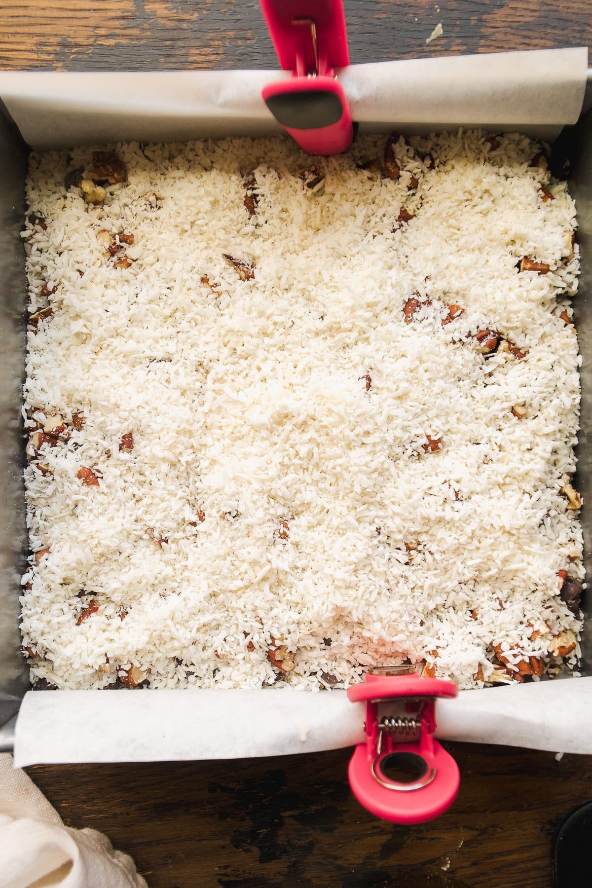 A square baking pan lined with parchment paper and pink clips, filled with an even layer of shredded coconut and chopped nuts&mdash;perfect for making vegan 7 layer bars, ready to be baked.