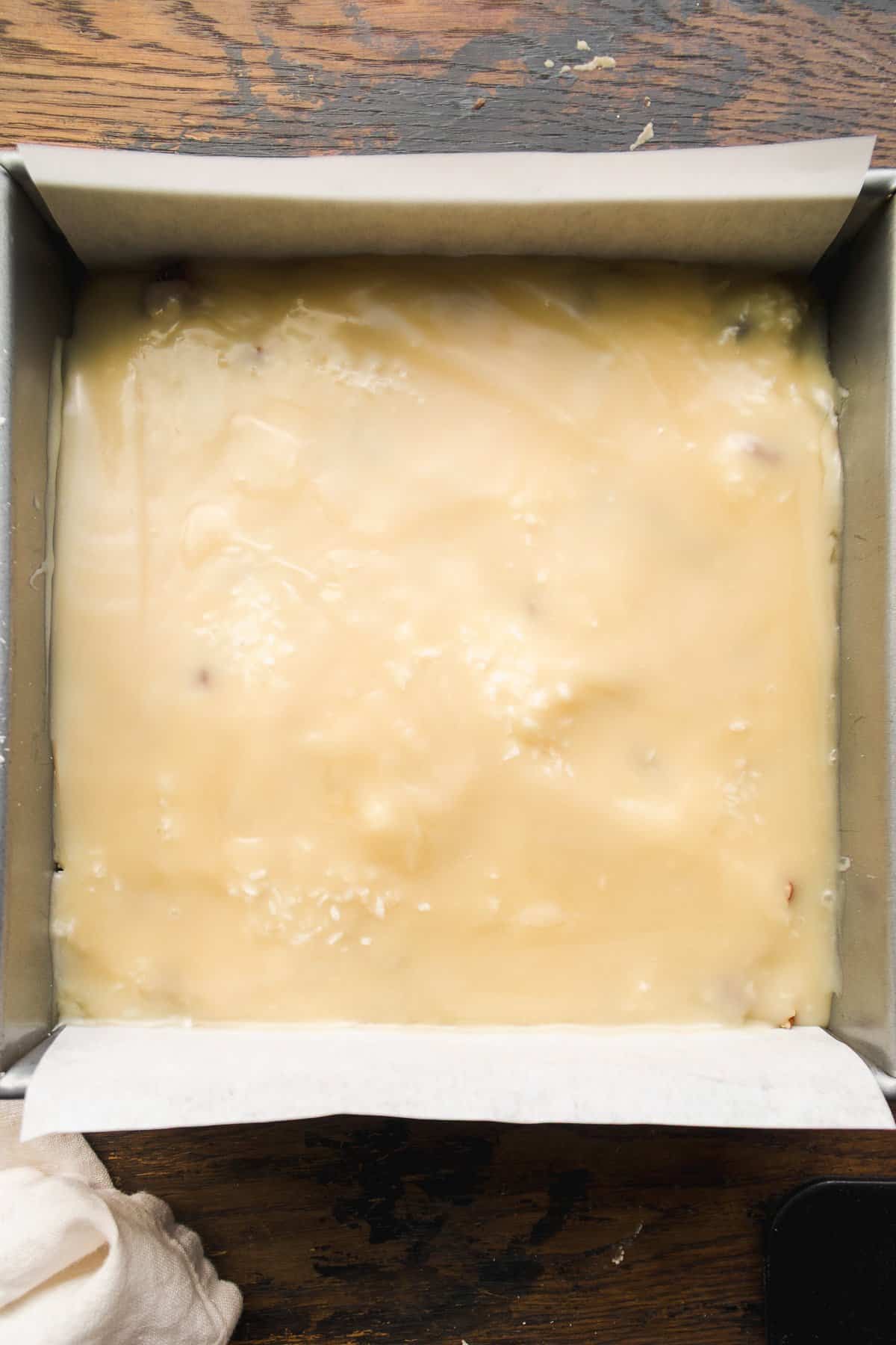 A square baking pan lined with parchment paper, filled with a smooth, pale yellow mixture for vegan 7 layer bars, rests on a wooden surface.