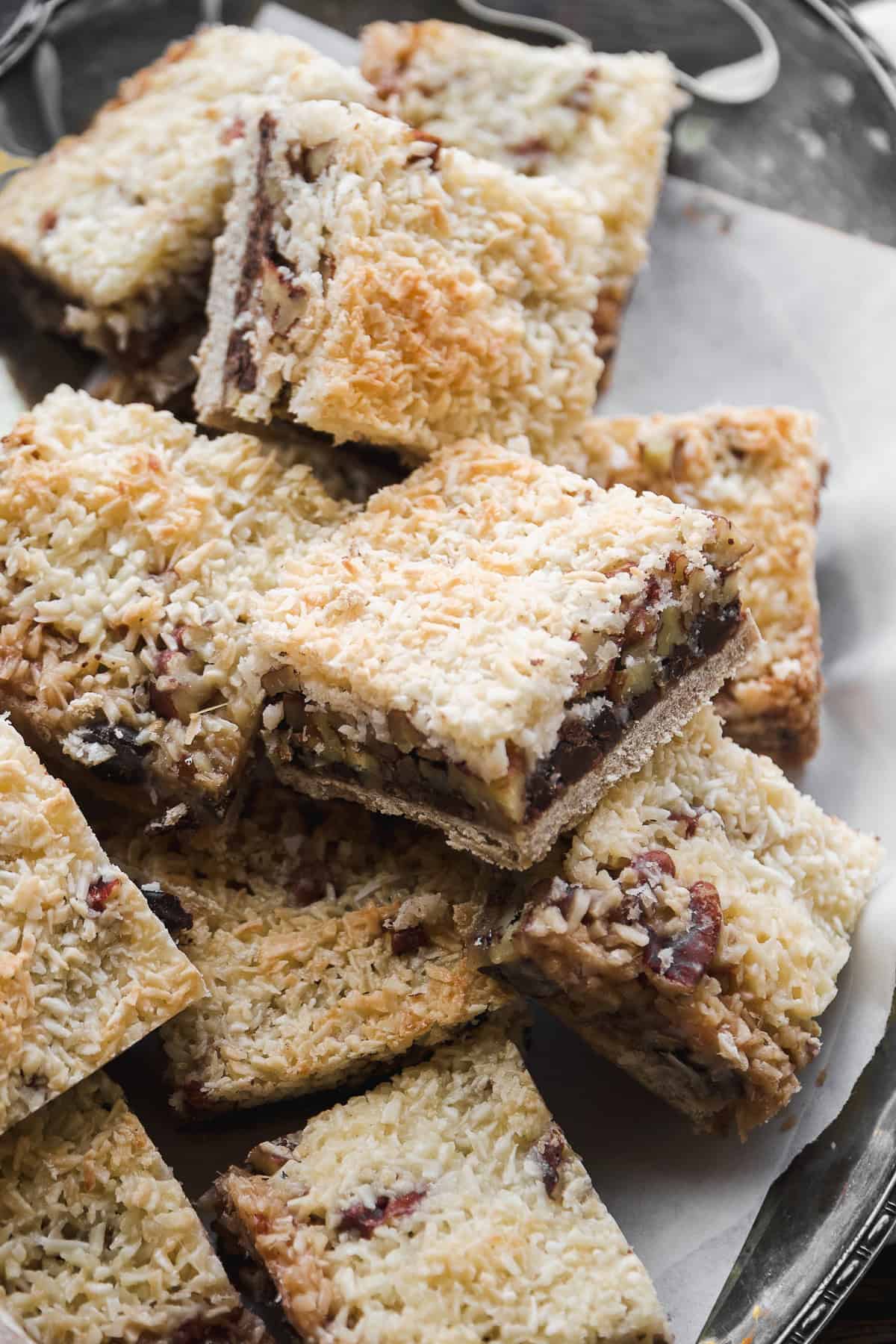 A close-up of a tray filled with vegan 7 layer bars, topped with coconut and featuring layers of baked crust, chocolate, and dried fruit. The bars are cut into squares and stacked on parchment paper.