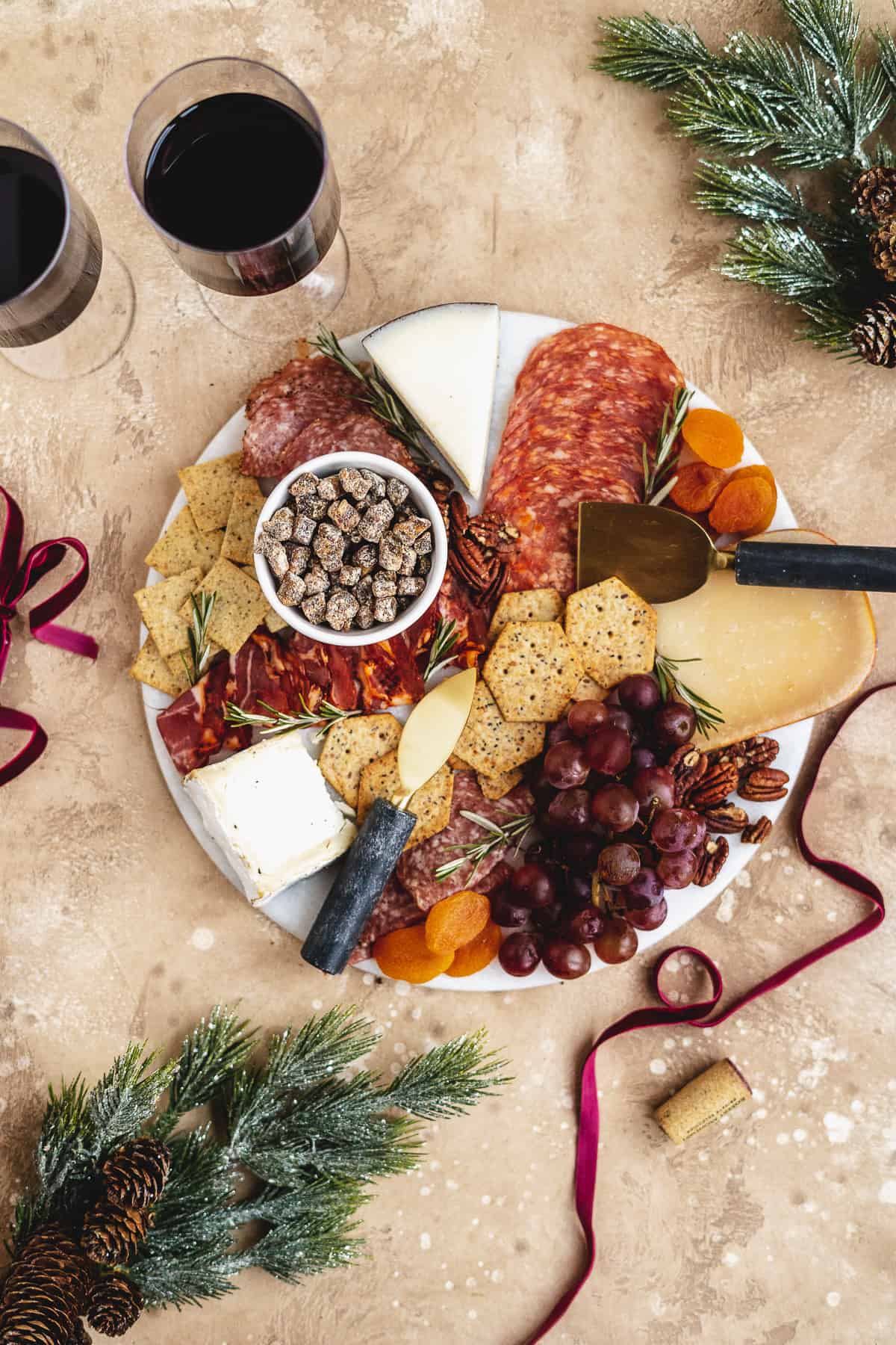 Overhead photo of round white marble slab covered with grapes, pecans, dried fruits, and assorted meats and cheeses.  Two glasses of red wine and gold cheese knives sit nearby.  