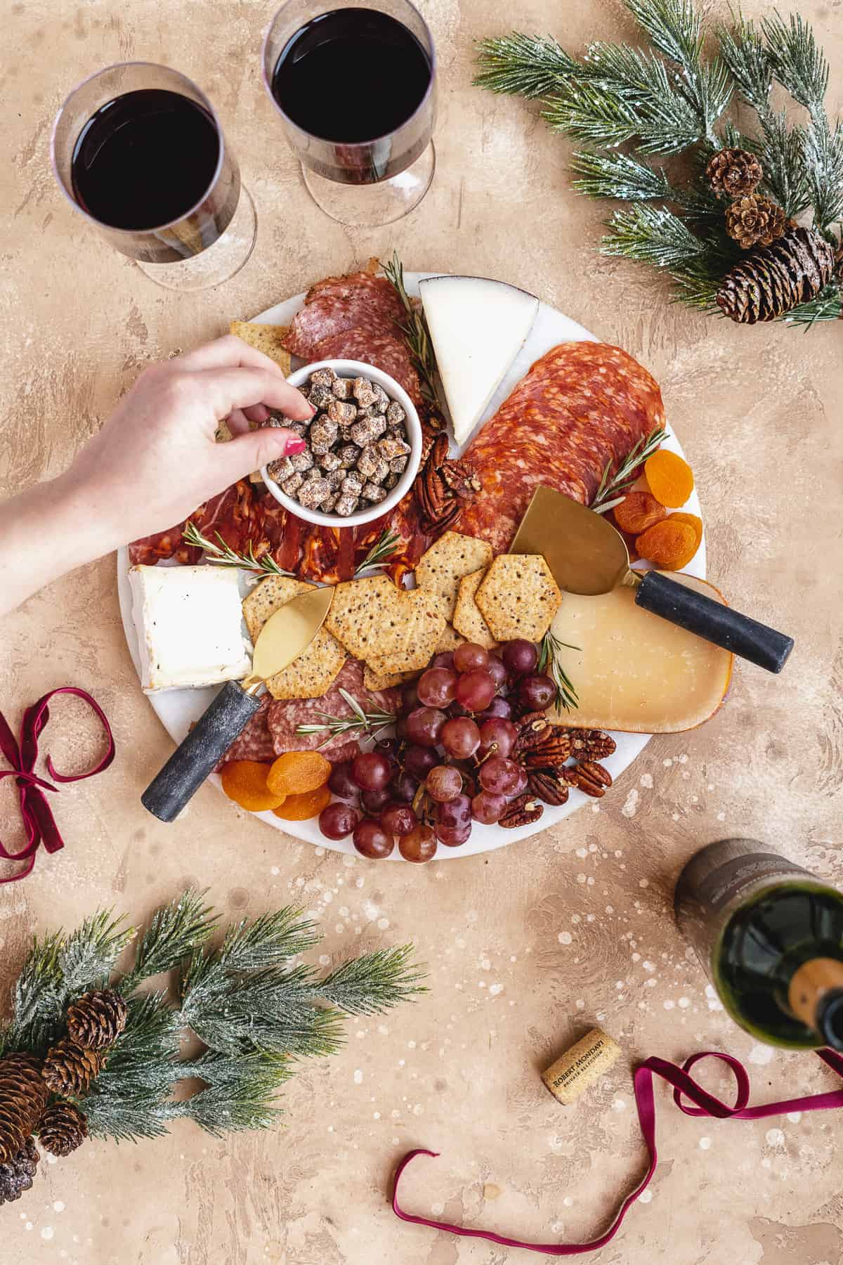 Overhead photo of round white marble cake platter filled with assorted meats, cheeses, fruits, crackers and nuts.  Two glasses of red wine sit nearby.  Hand is reaching to pick up some nuts from the charcuterie board.  