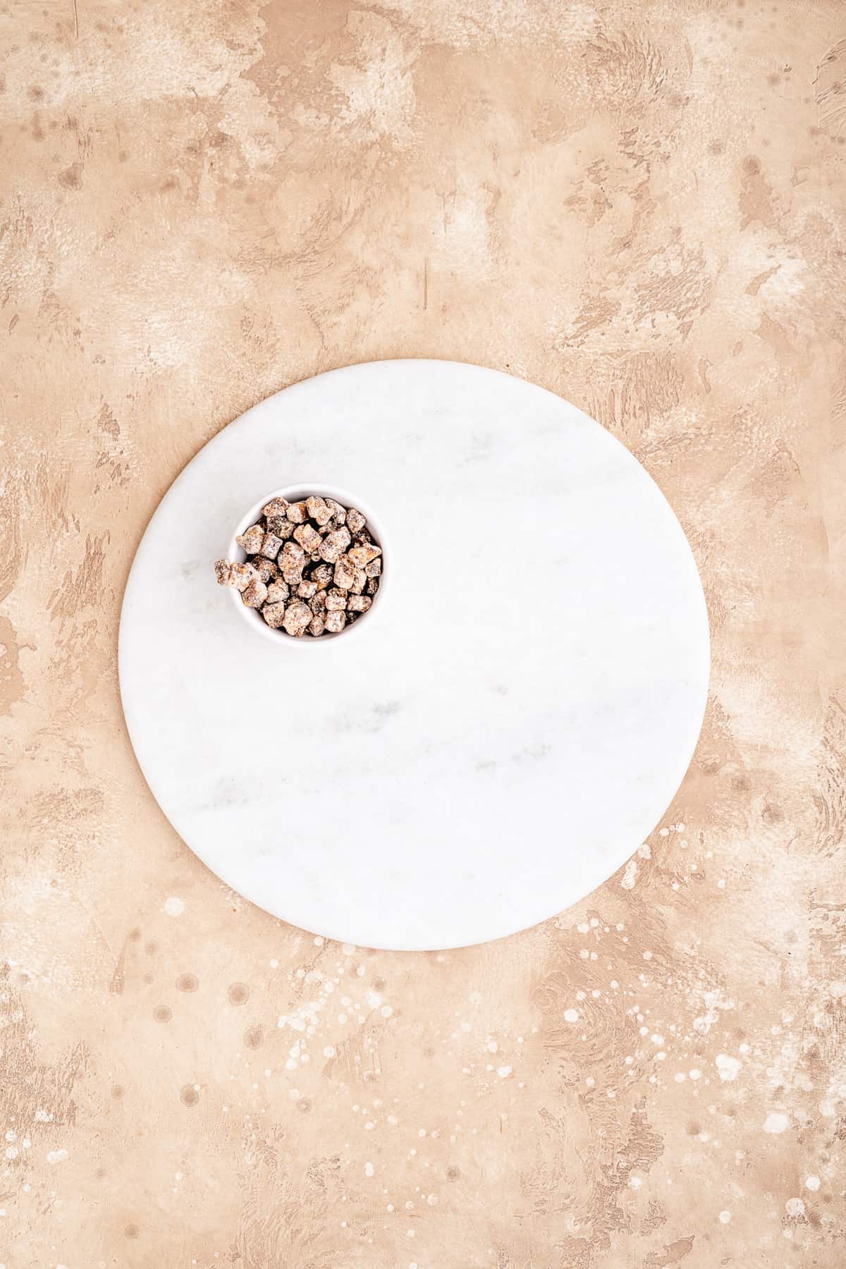 Overhead photo of white marble cake platter with small white bowl of nuts placed on the platter to begin building the charcuterie board.