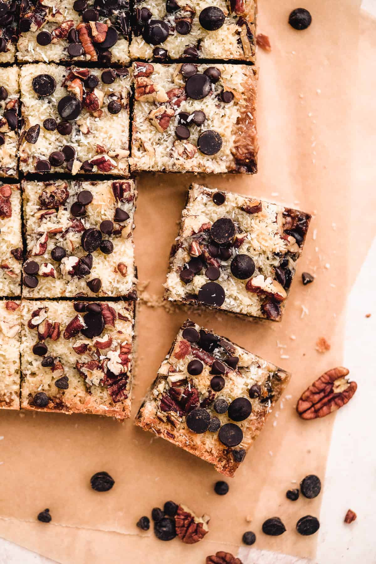 Overhead view of freshly baked Toasted Oat Seven Layer Magic Cookie Bars cut into squares.  Pecans and chocolate chips are sprinkled around.  