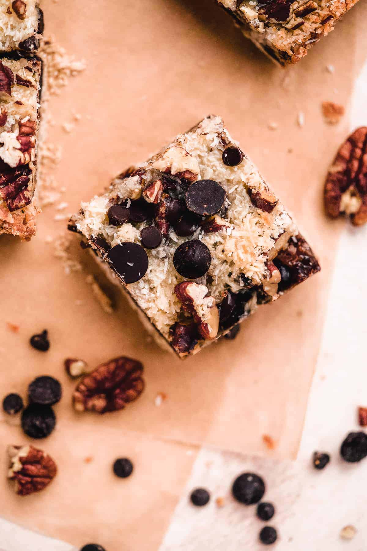 Closeup overhead view of a single Toasted Oat Seven Layer Magic Cookie Bar on parchment paper.  Pecans and chocolate chips are sprinkled around.  