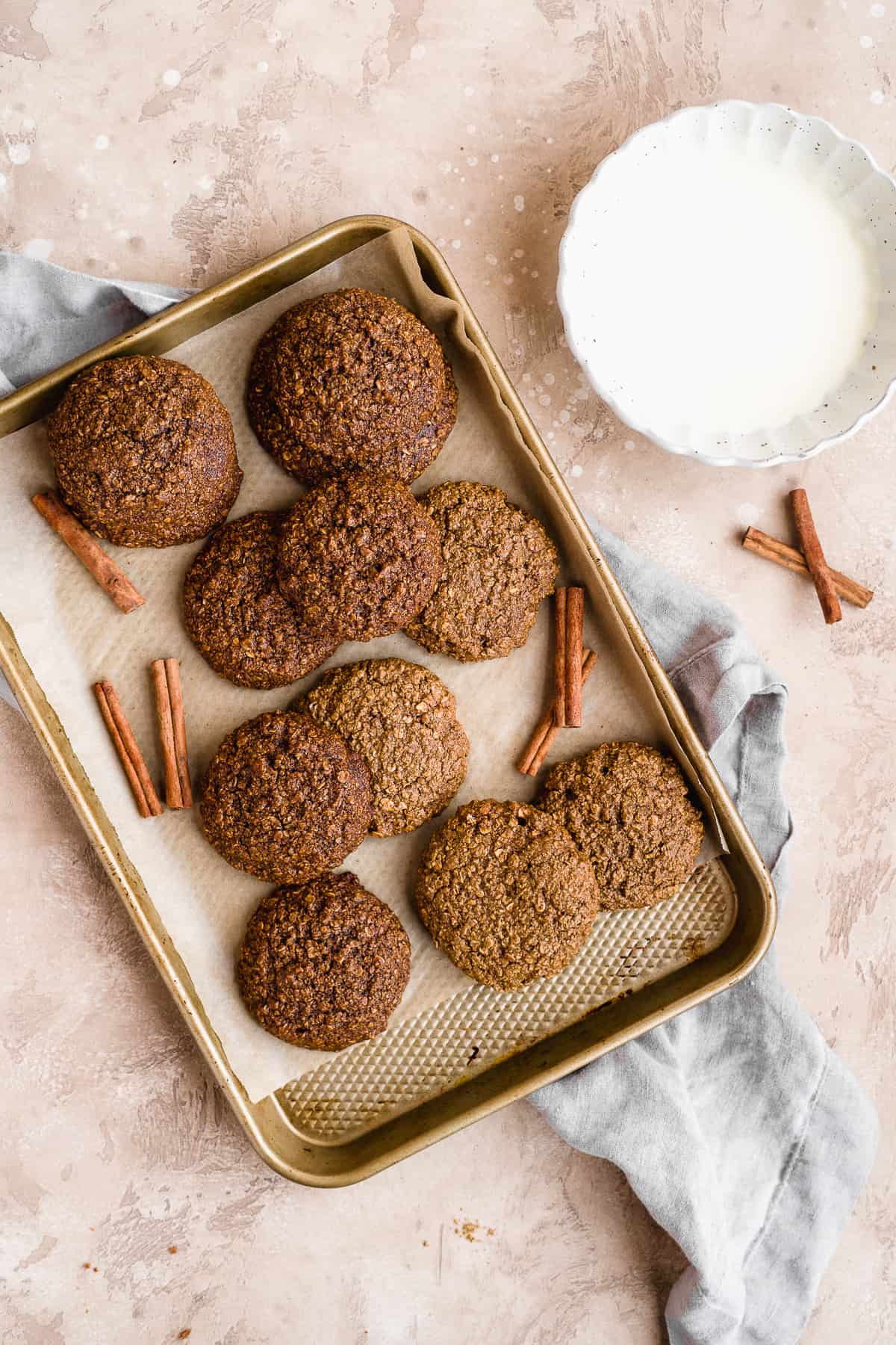 Gold baking sheet with oatmeal gingerbread cookies fresh out of the oven waiting to be dipped into the white chocolate mix.  White scalloped bowl of melted chocolate sits nearby.  Cinnamon sticks are scattered around.   