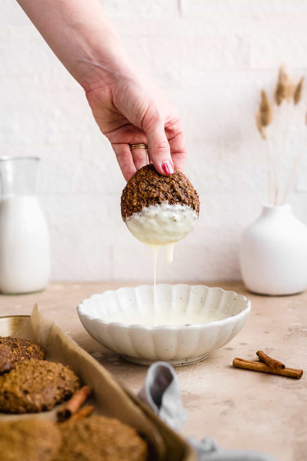 White scalloped bowl with decadent white chocolate.  View of hand holding a yummy oatmeal gingerbread cookie as it's dipped into the white chocolate and chocolate drips off the cookie.  Cinnamon sticks and additional cookies sit nearby.  