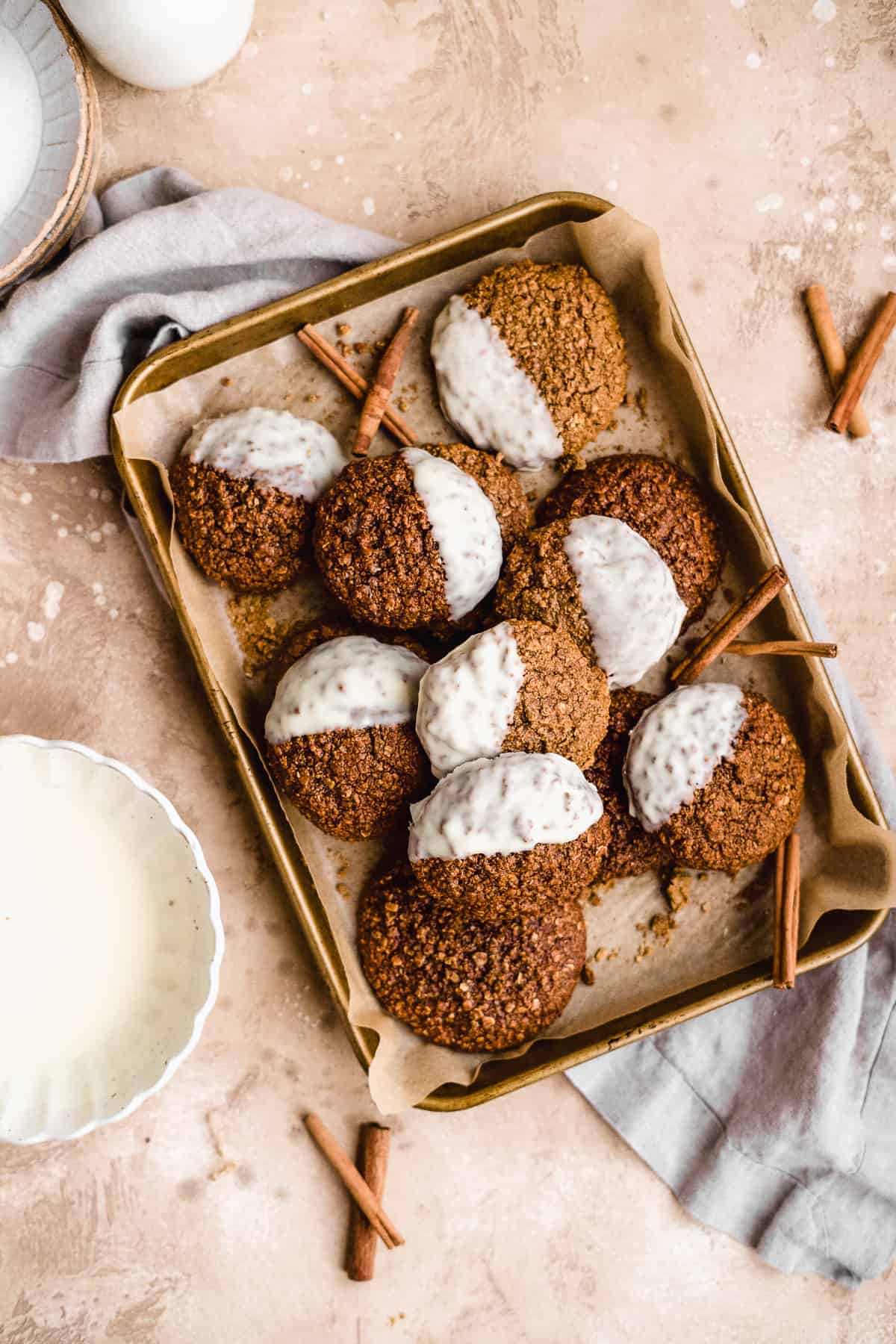 Gold baking sheet with parchment paper with freshly baked oatmeal gingerbread cookies dipped in white chocolate.  Cinnamon sticks are scattered nearby.  A white scalloped bowl with white chocolate sits next to the tray.  