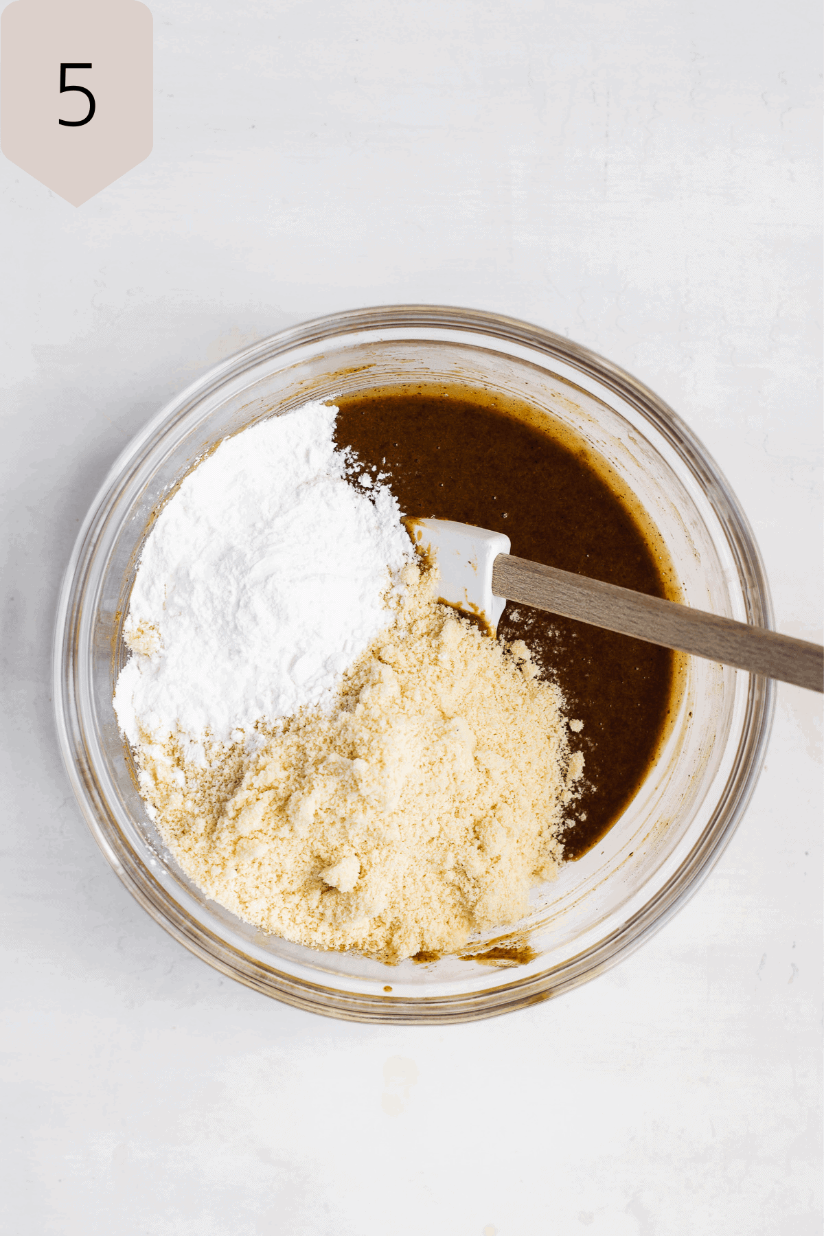 Glass mixing bowl with flour about to be mixed into cookie batter on a white surface.