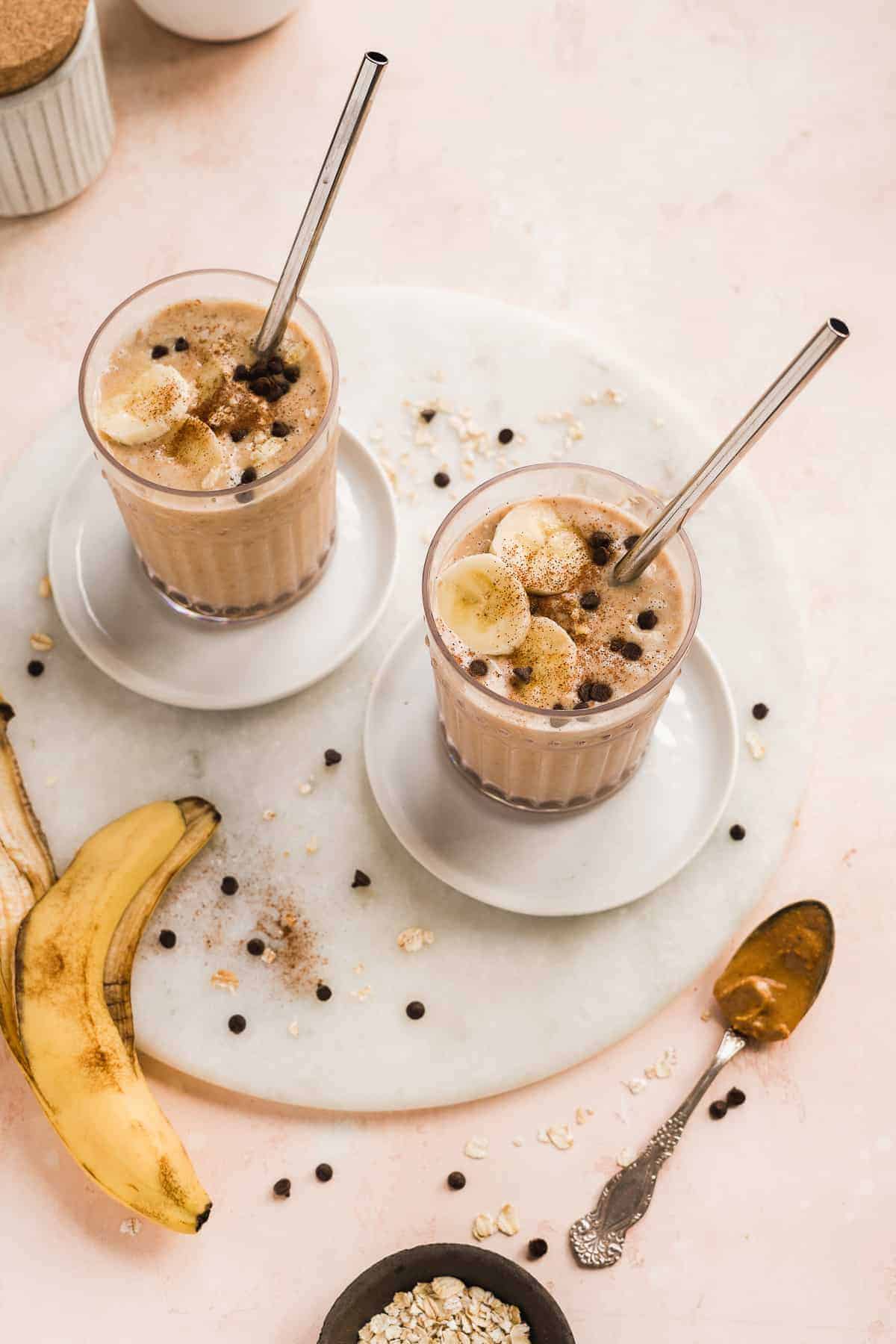 Two smoothie glasses with toppings and a straw on a marble platter with a spoon of nut butter.